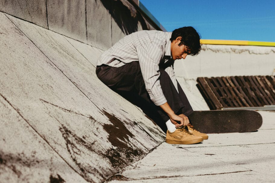 Een jonge man zit op een betonnen helling en strikt de veters van zijn bruine sneakers naast zijn skateboard