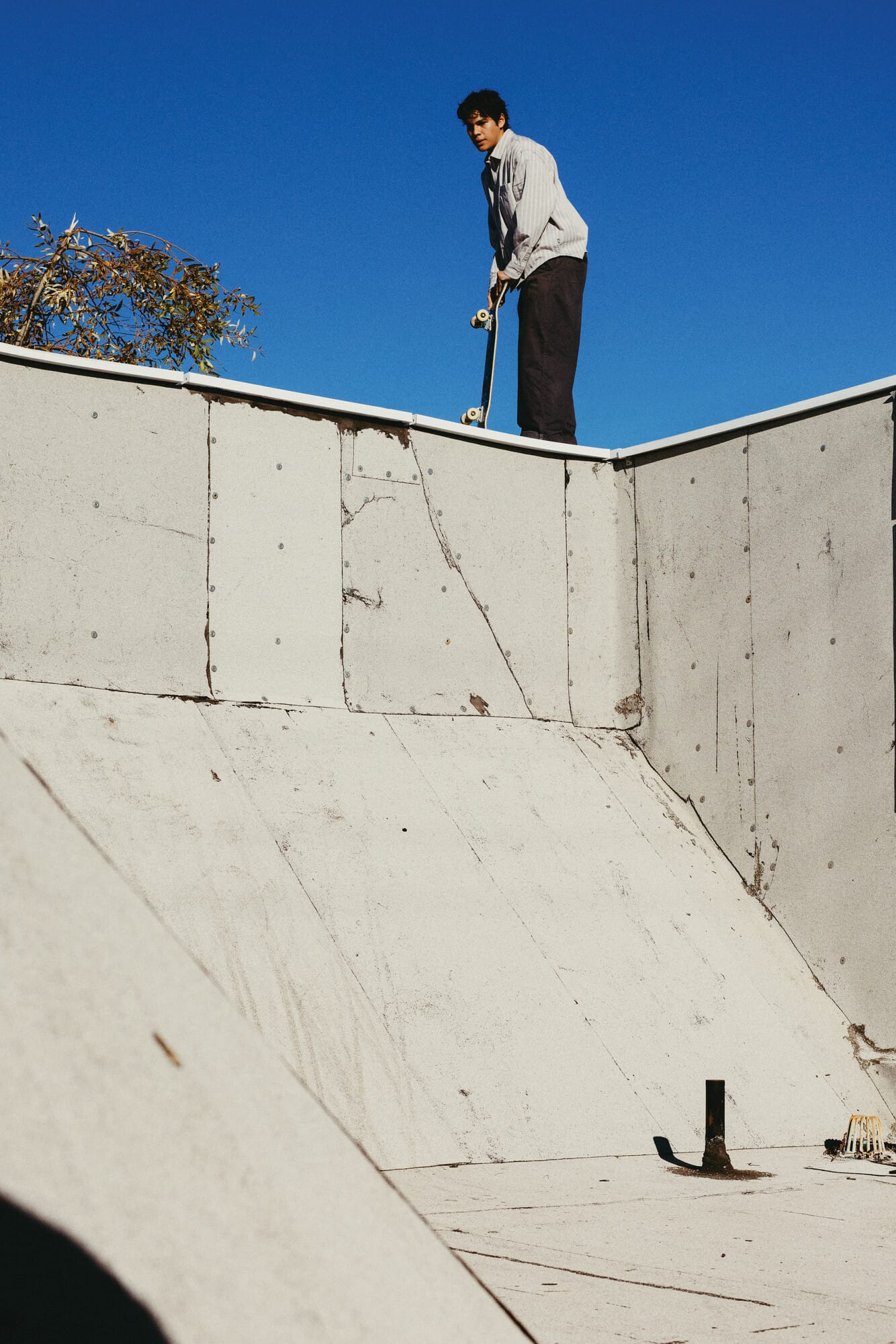 Een skateboarder staat met zijn skateboard bovenaan de rand van een betonnen skatebaan tegen een strakblauwe lucht