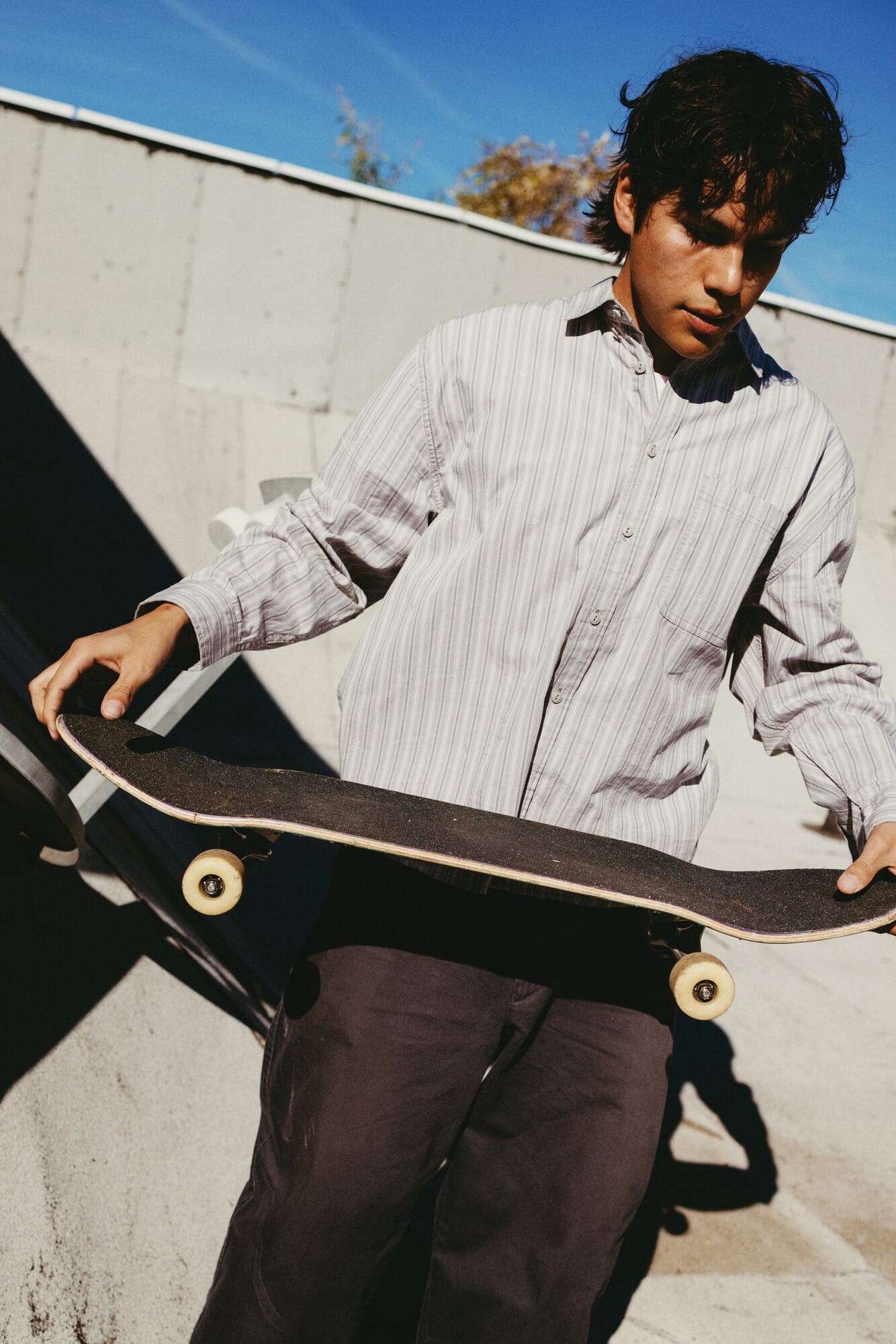 Een jonge man in een gestreept overhemd houdt een skateboard met beide handen vast in de zon