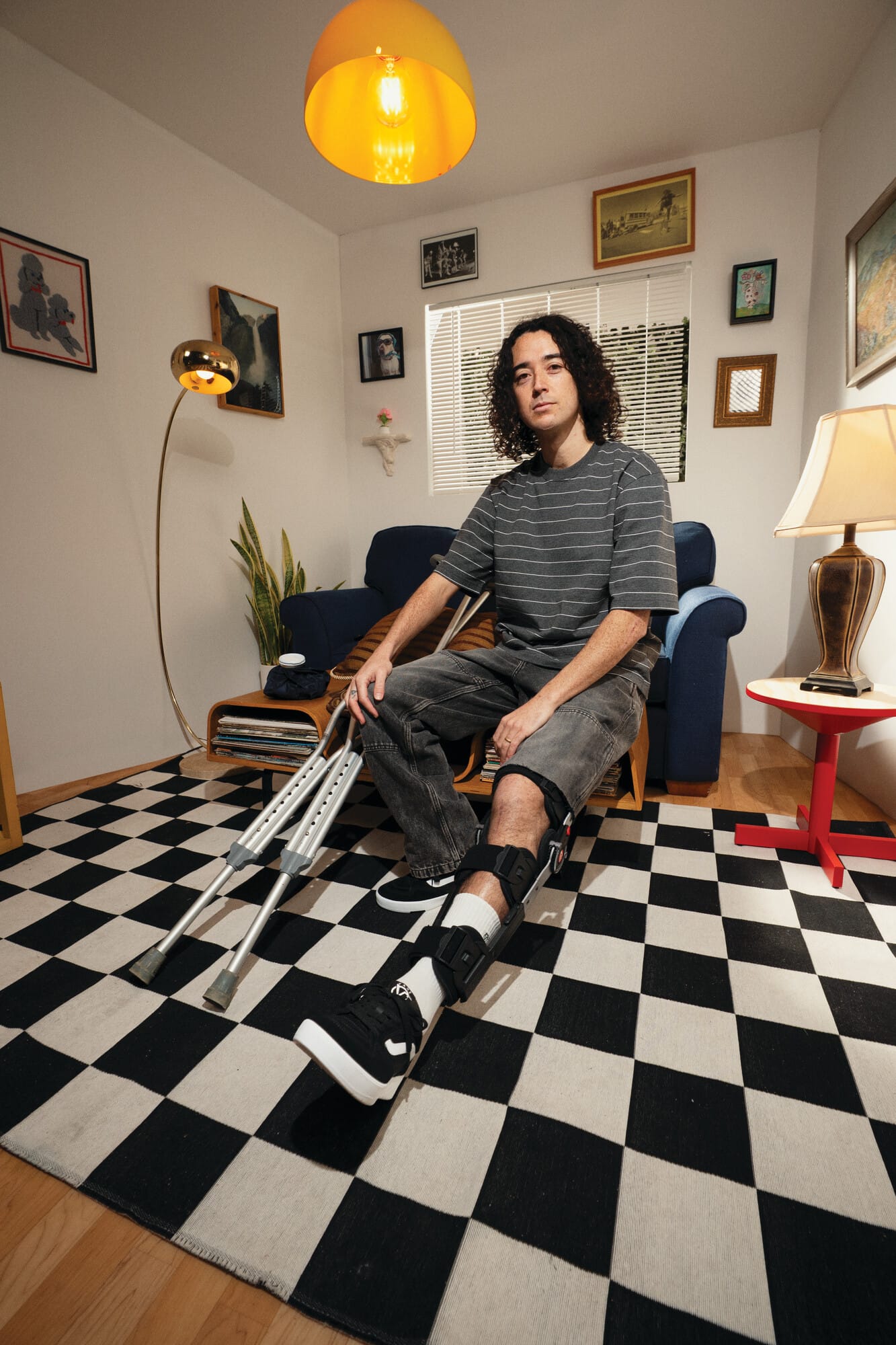 Een man met een beenbrace zit in een woonkamer met krukken naast zich op een geblokt tapijt