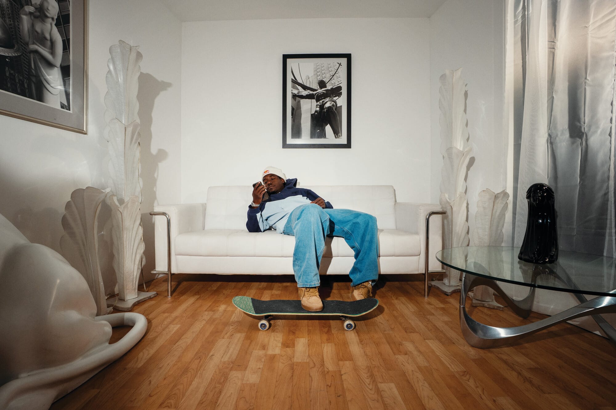 Een man zit op een witte bank en kijkt op zijn telefoon, met één voet rustend op een skateboard op de vloer