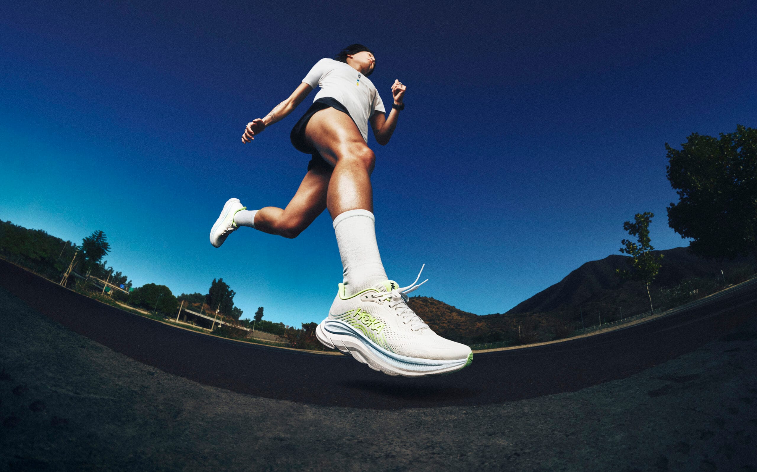 Hardloper in actie op een weg, gezien vanuit een laag perspectief met de focus op witte Hoka hardloopschoenen model Clifton 11