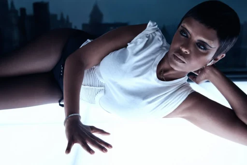 Zangeres Kelela met kort haar en een wit T-shirt ligt op een verlicht oppervlak tegen een donkere stadsachtergrond