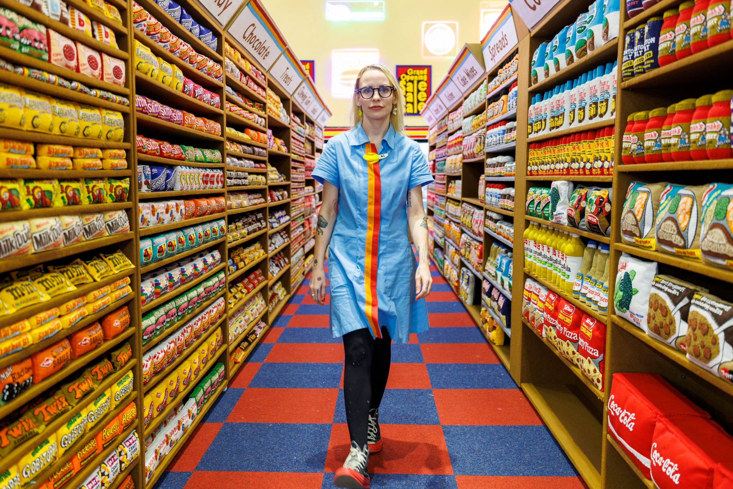 Kunstenaar Lucy Sparrow in een blauwe werkjas loopt door een gangpad van haar vilten supermarkt-installatie