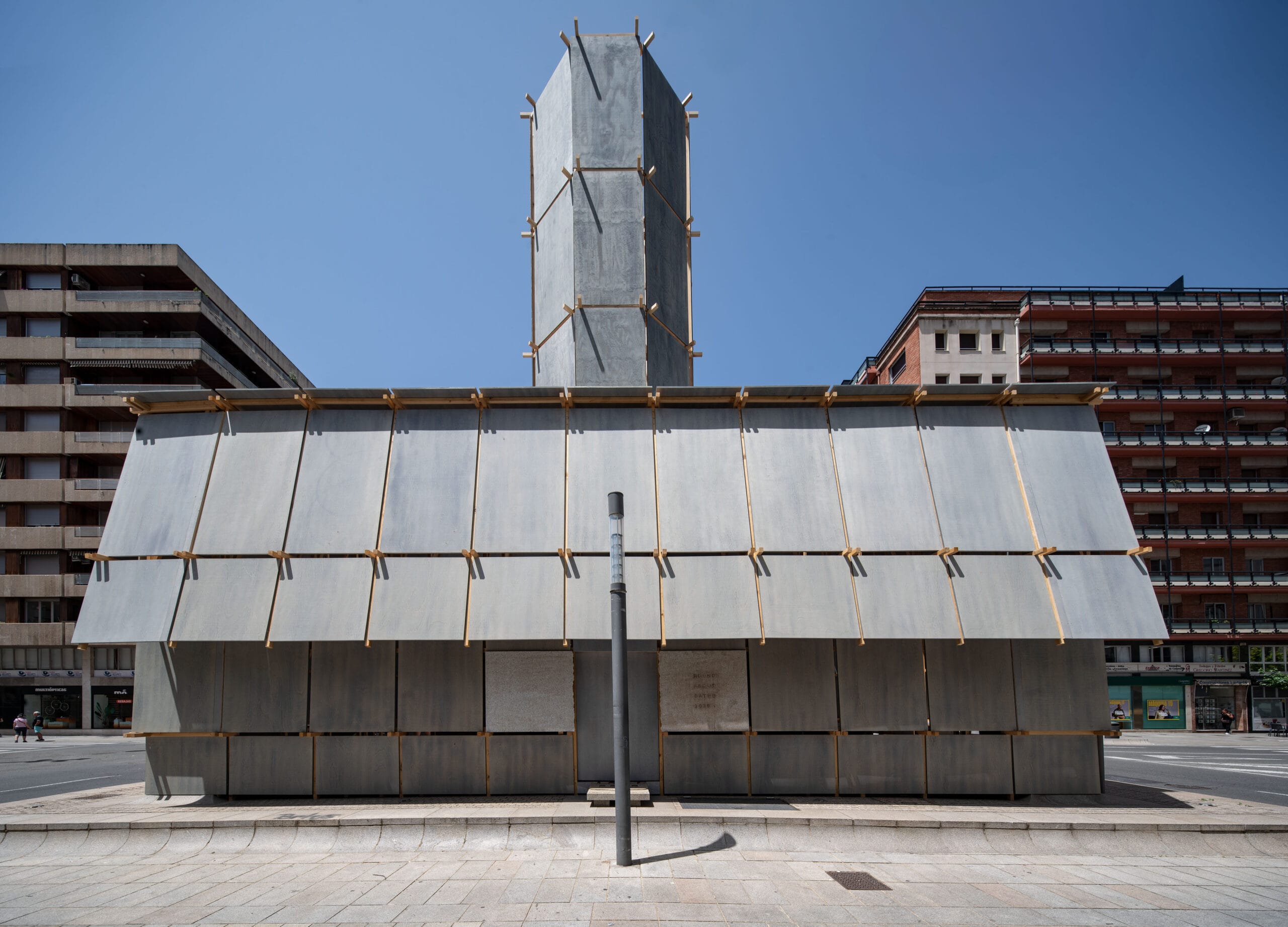 De Round About Baths installatie: een houten constructie met grijze panelen en een hoge toren op een rotonde in Logroño