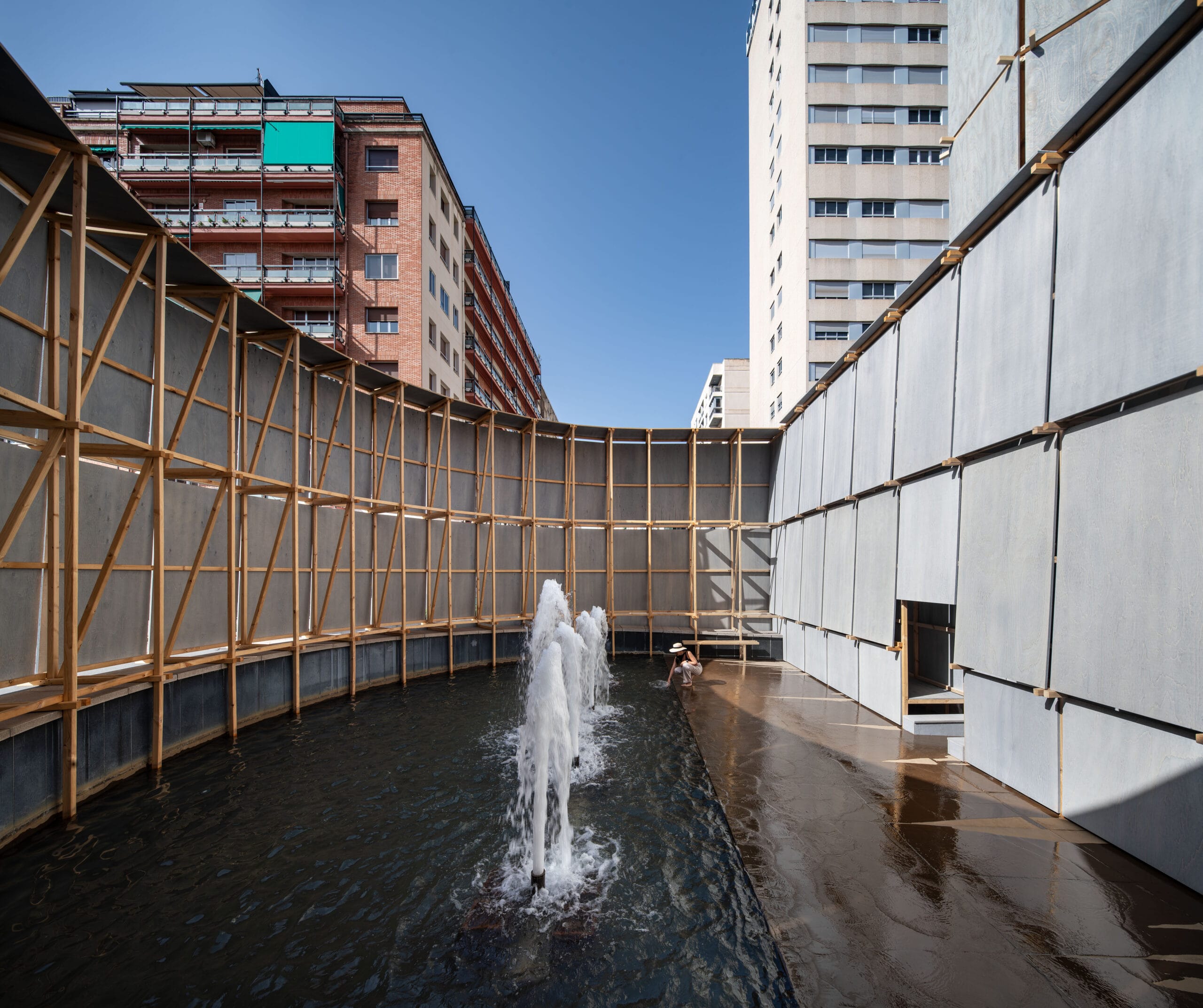 Een houten constructie met hoge wanden rondom een fontein op een rotonde, die dient als tijdelijk openbaar badhuis