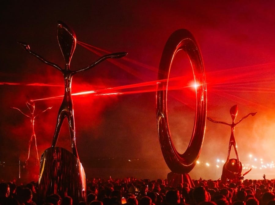 Grote abstracte menselijke sculpturen en een cirkel op een festivalterrein, verlicht door felrode lasers en rook