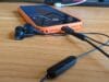 Een smartphone in een oranje hoesje op een houten tafel, verbonden met zwarte bedrade oortjes die muziek afspelen