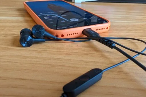 Een smartphone in een oranje hoesje op een houten tafel, verbonden met zwarte bedrade oortjes die muziek afspelen