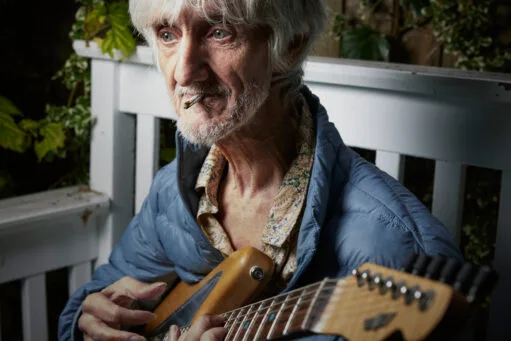 Oudere man met grijs haar en een sigaret in zijn mond speelt op een elektrische gitaar
