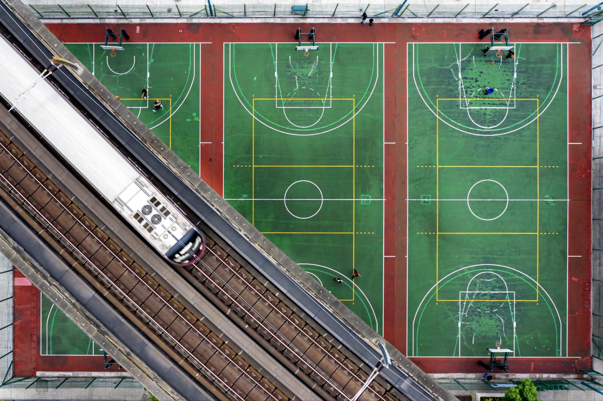 Bovenaanzicht van drie felgroene basketbalvelden met rode randen waar diagonaal een metrolijn overheen loopt