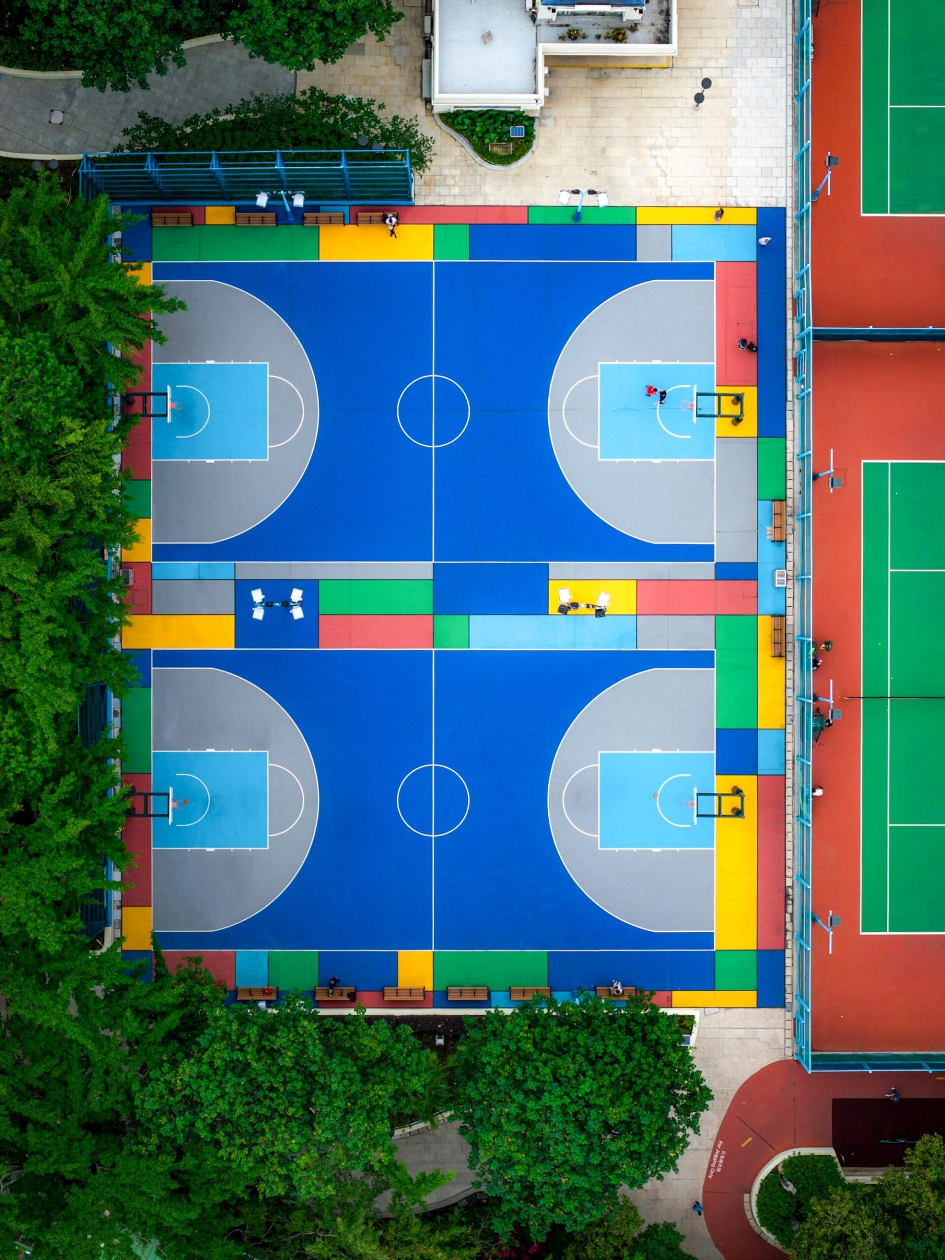 Bovenaanzicht van twee blauwe basketbalvelden met kleurrijke geometrische patronen, omringd door bomen en tennisbanen