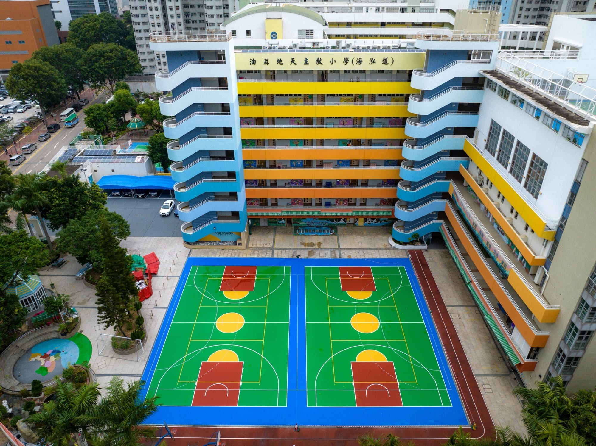 Bovenaanzicht van een felgekleurd blauw en groen basketbalveld naast een basisschool met kleurrijke gevels in Hong Kong