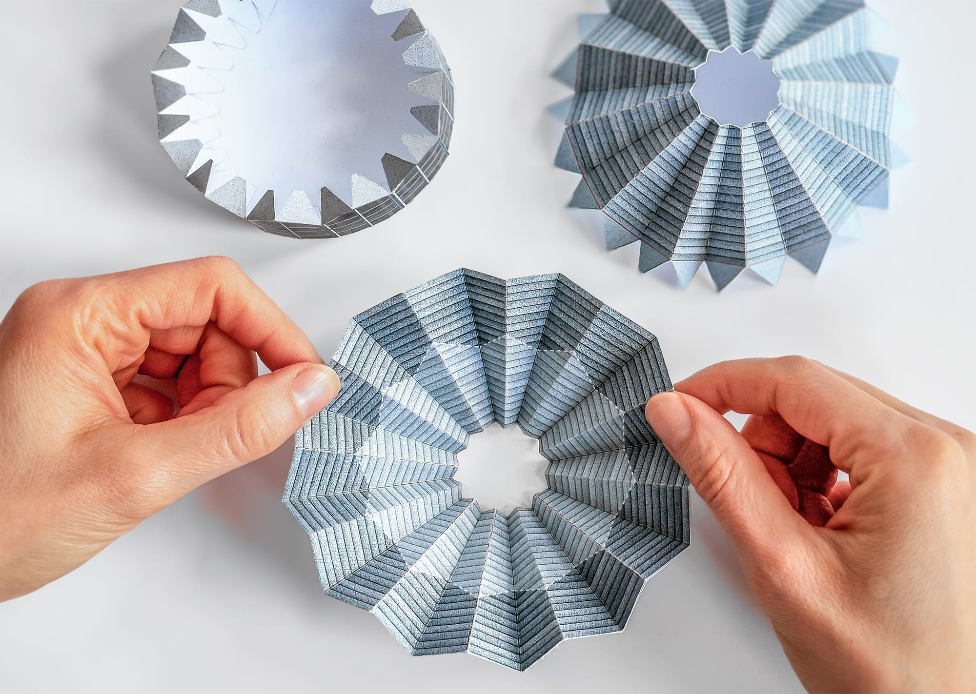Handen die een geometrisch gevouwen papieren object vasthouden naast twee andere gevouwen papieren vormen op een witte tafel