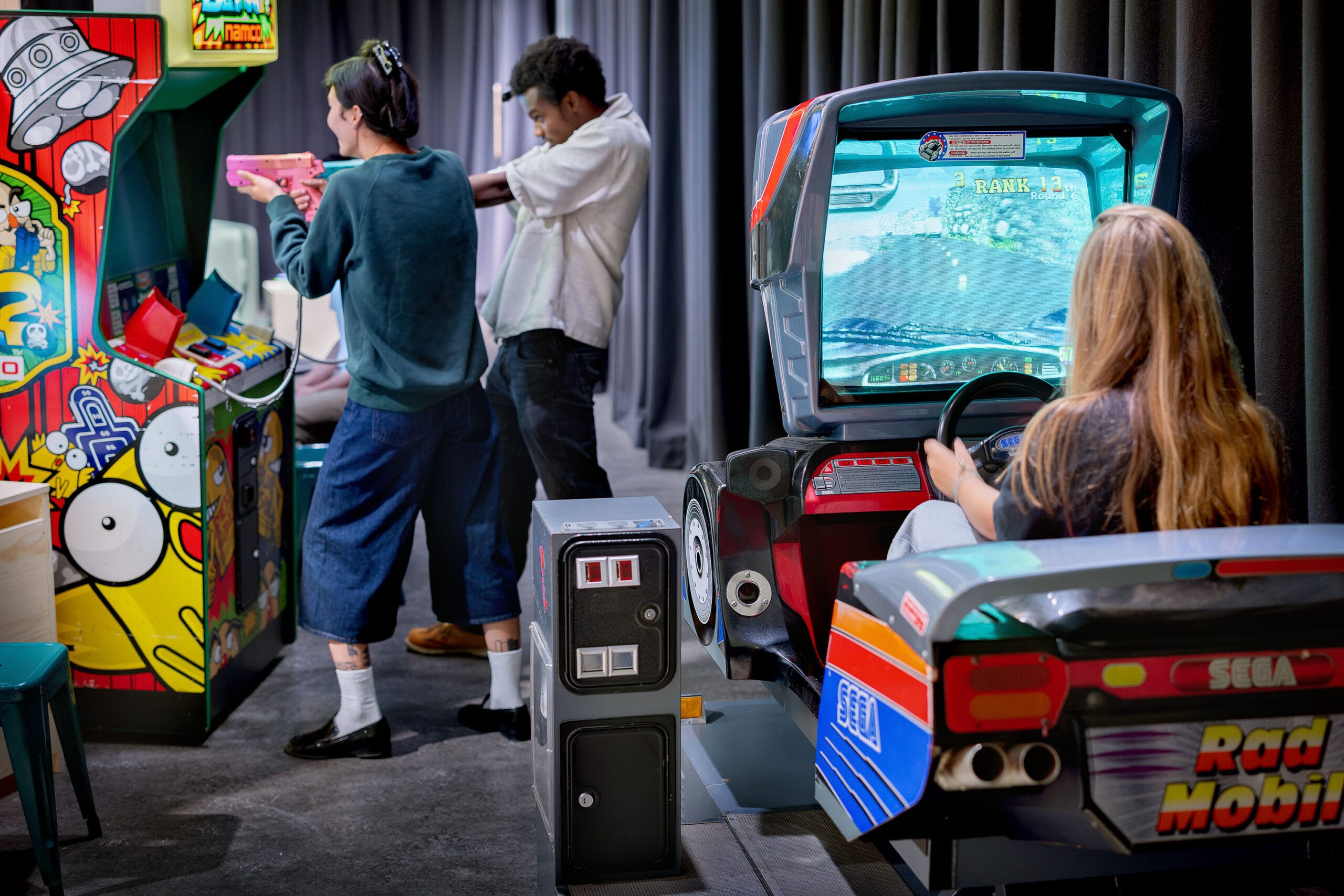 Mensen spelen op verschillende retro arcadekasten, waaronder een schietspel en een racespel van Sega