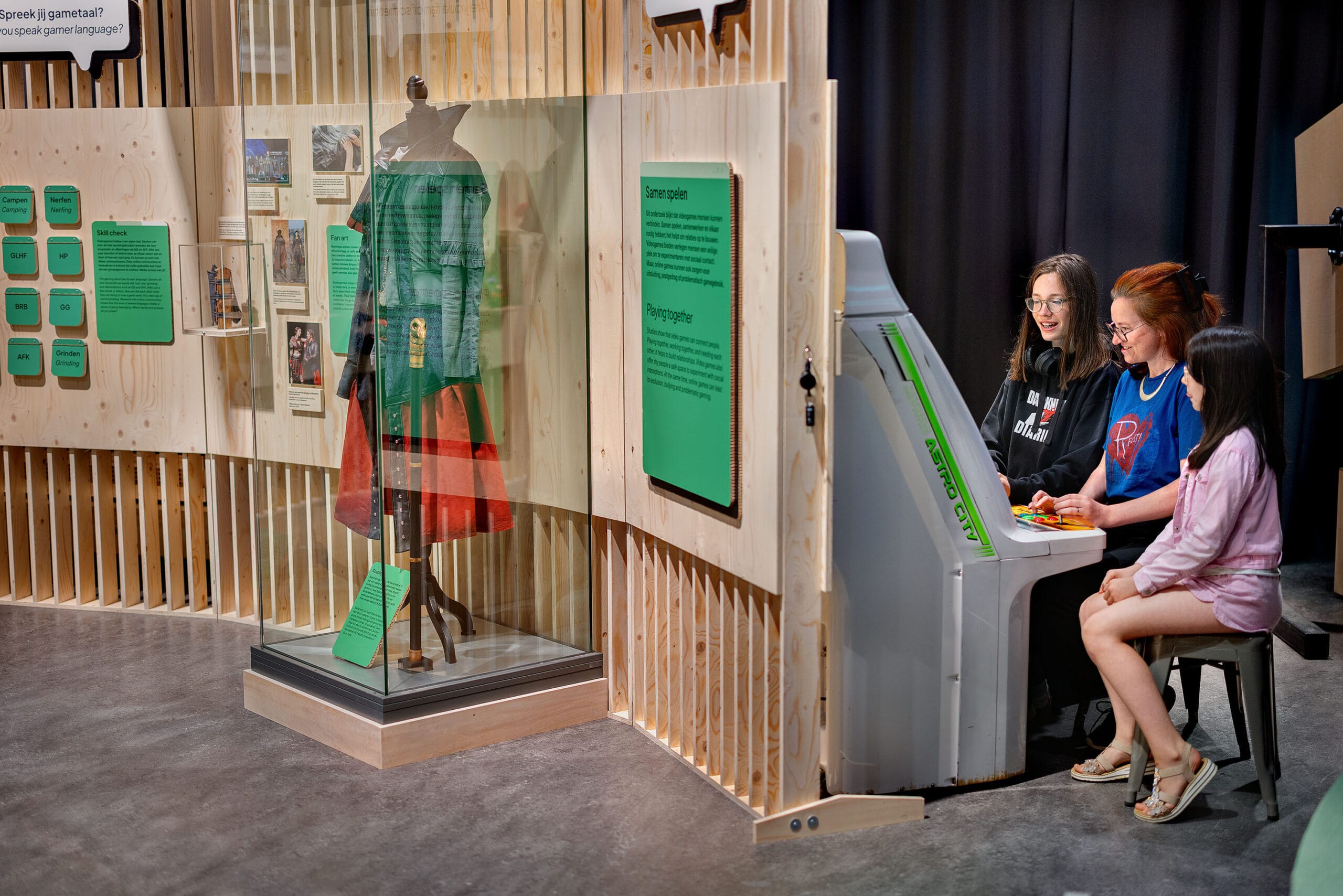 Drie bezoekers spelen samen op een arcadekast in een museum over gaming, naast een vitrine met een kostuum