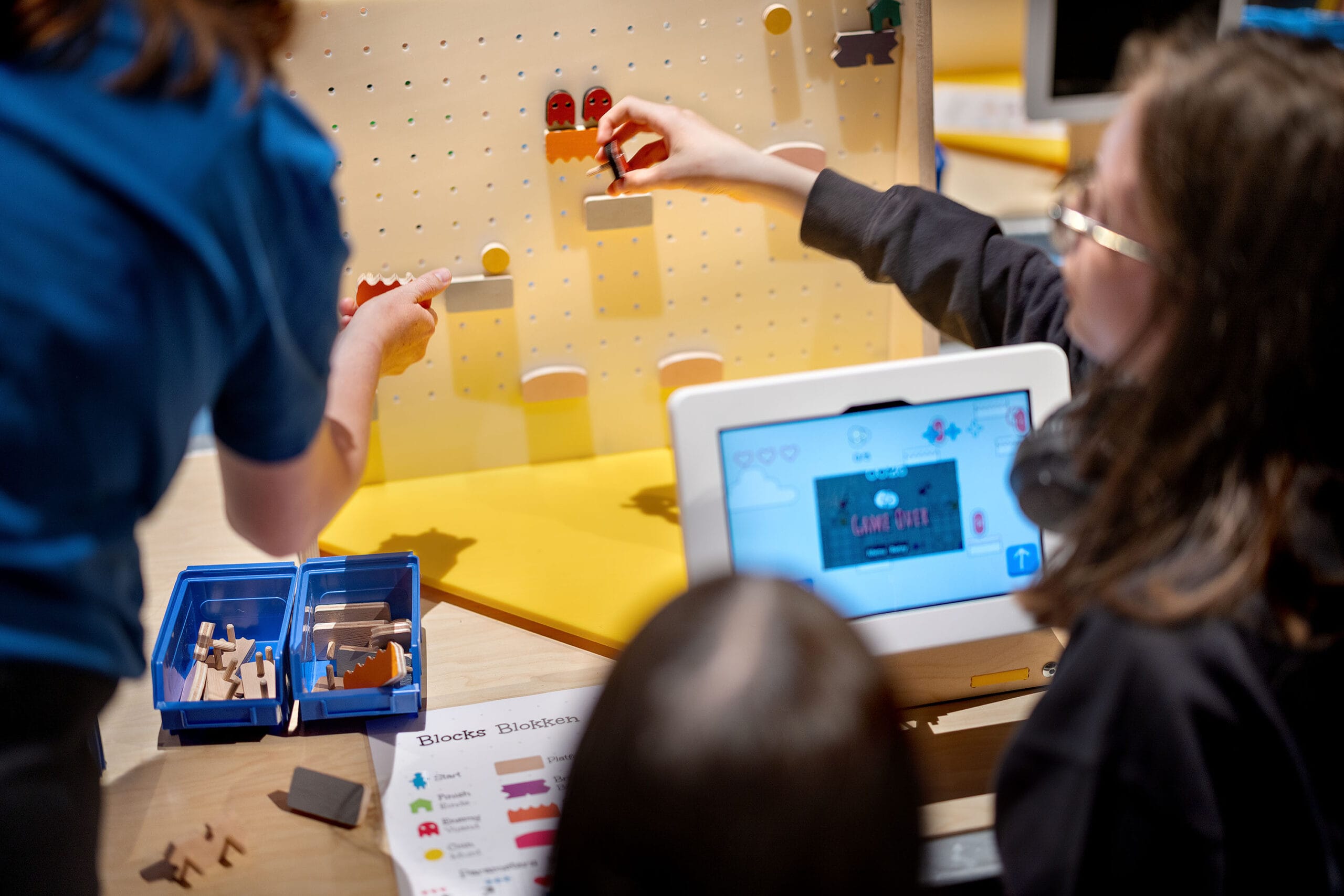 Twee personen plaatsen houten blokken op een pegboard om een fysiek level te bouwen voor een digitaal spel op een tablet