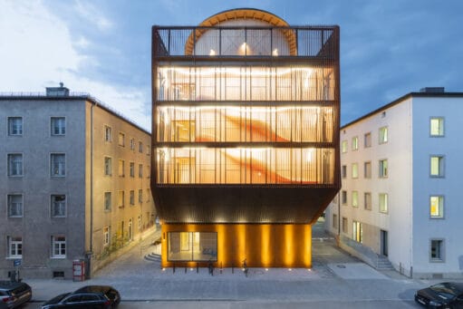 Modern verlicht gebouw met een houten lattengevel tussen twee traditionele stadsgebouwen in de avond