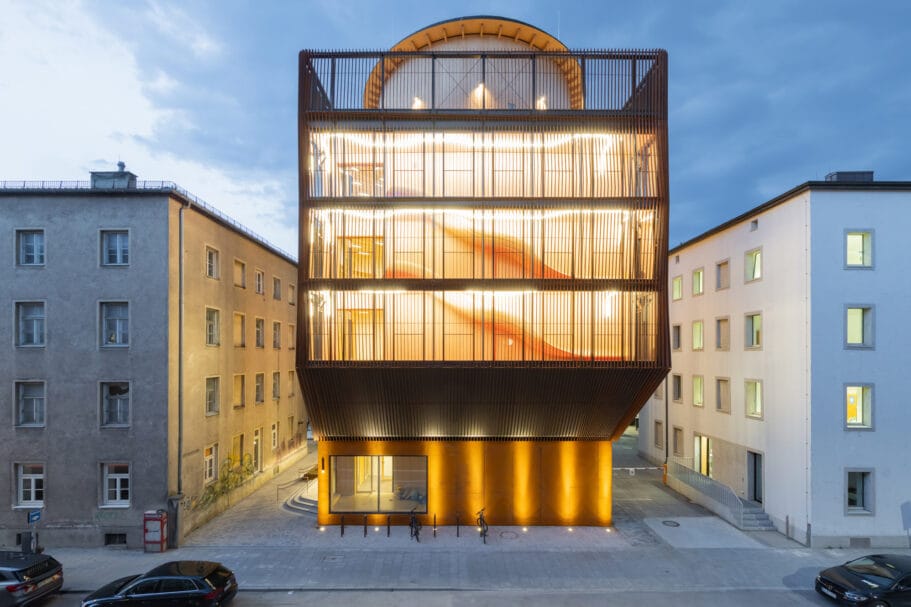 Modern verlicht gebouw met een houten lattengevel tussen twee traditionele stadsgebouwen in de avond
