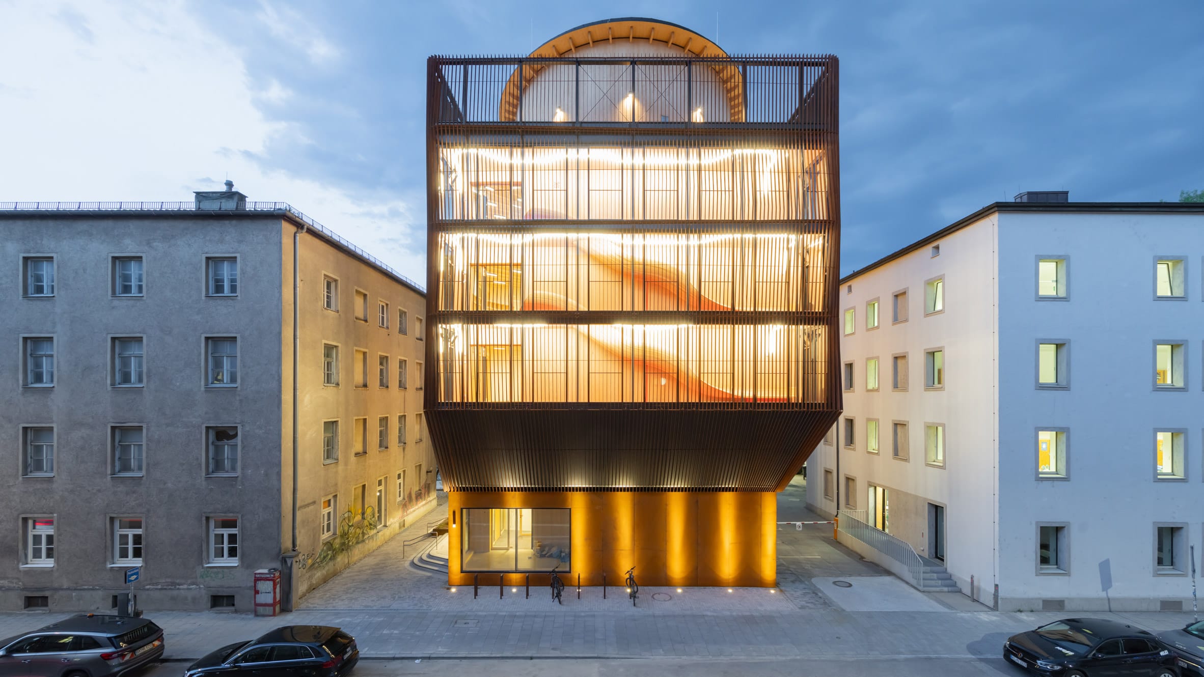 Modern verlicht gebouw met een houten lattengevel tussen twee traditionele stadsgebouwen in de avond