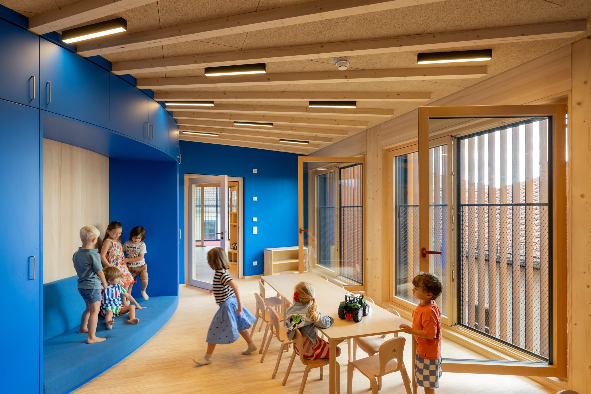Spelende kinderen in een moderne, lichte klasruimte met blauwe kasten, houten balkenplafond en grote ramen