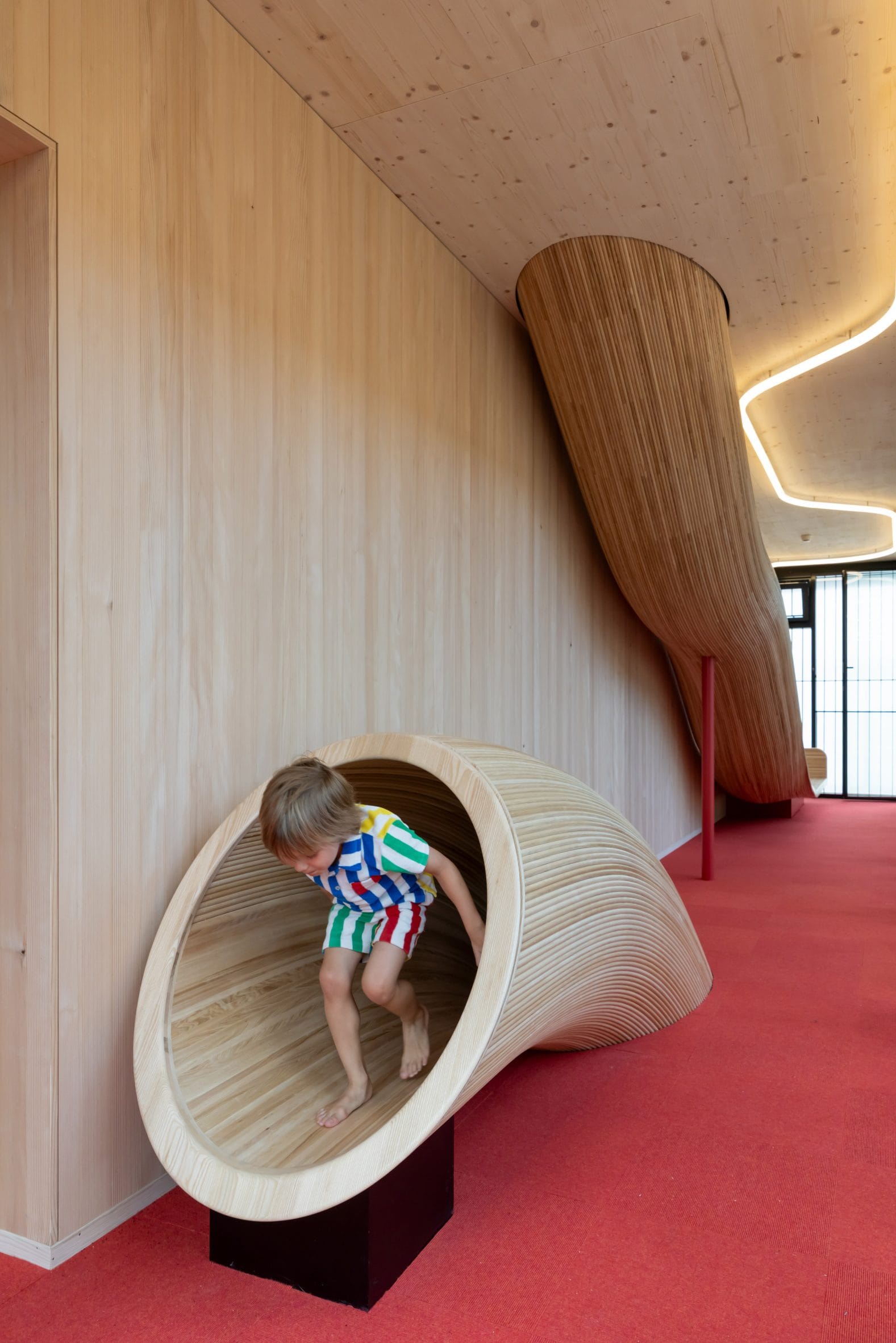 Een kind in een kleurrijk gestreept pakje kruipt uit een houten speeltunnel in een modern interieur met rode vloerbedekking