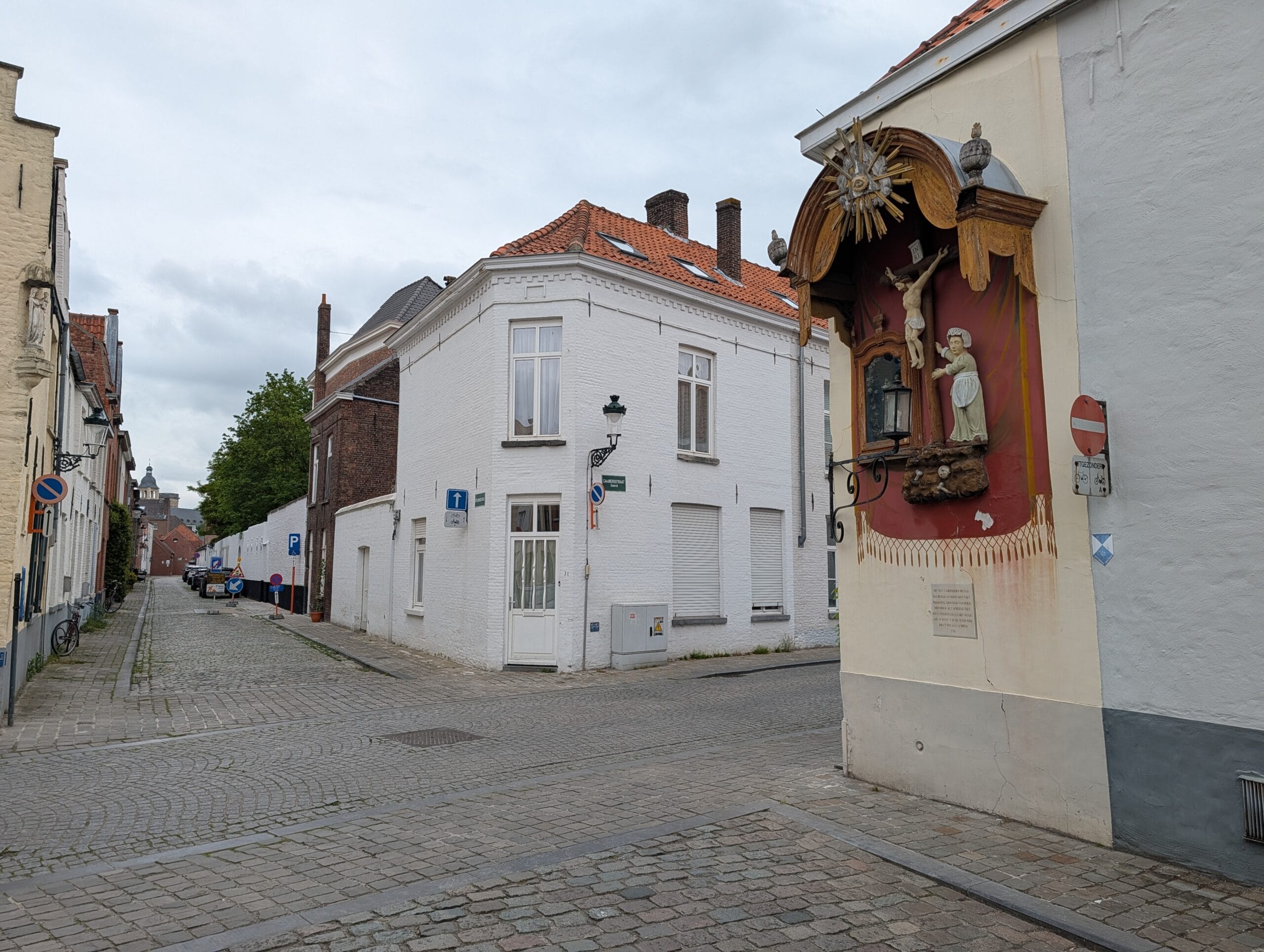 Een gevelkapel met een kruisbeeld aan een wit huis in een geplaveide straat in een historisch stadscentrum