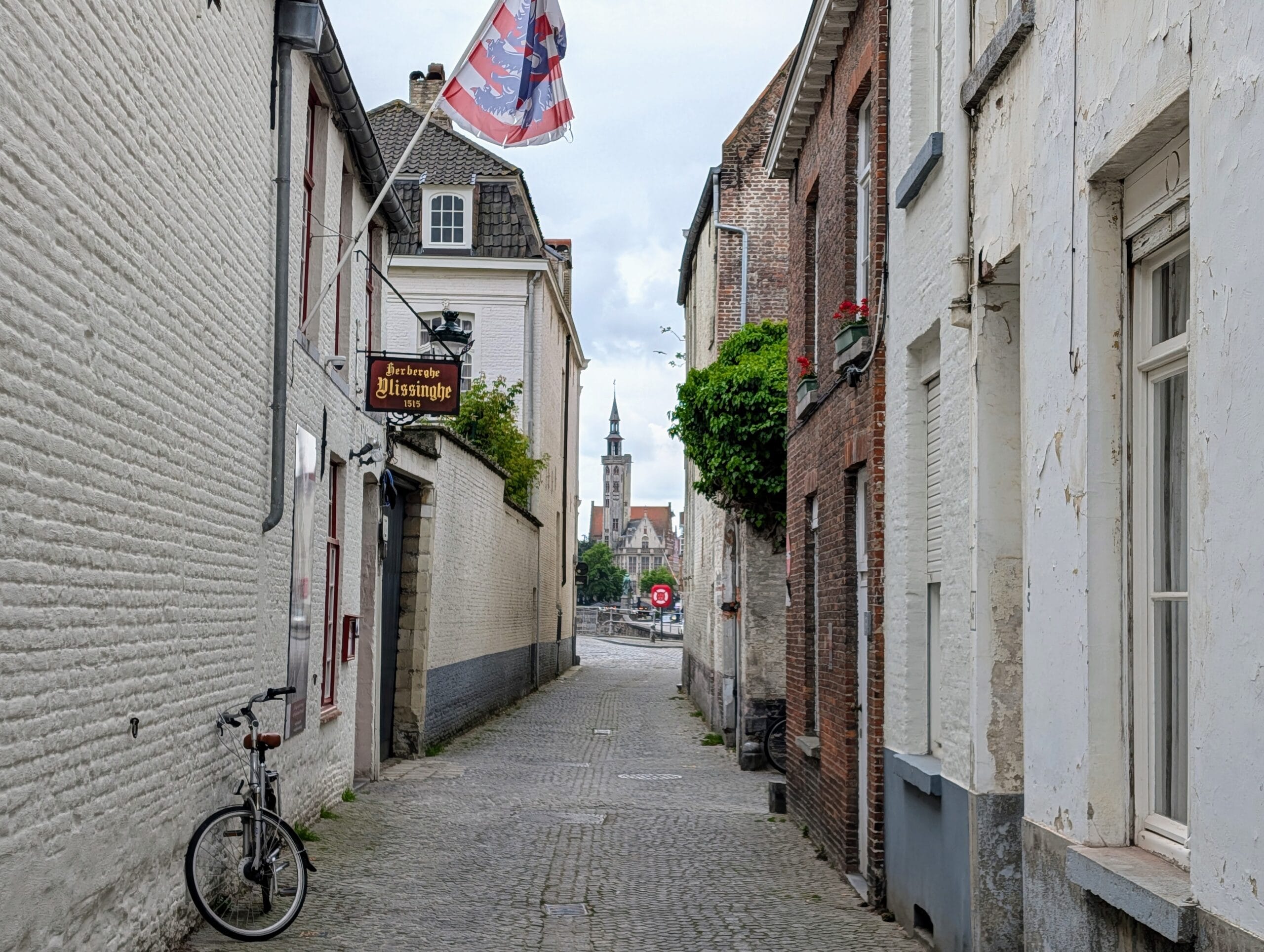 Smal kasseistraatje in Brugge met witte gevels, een fiets tegen de muur en de toren van het Poortersloge in de verte