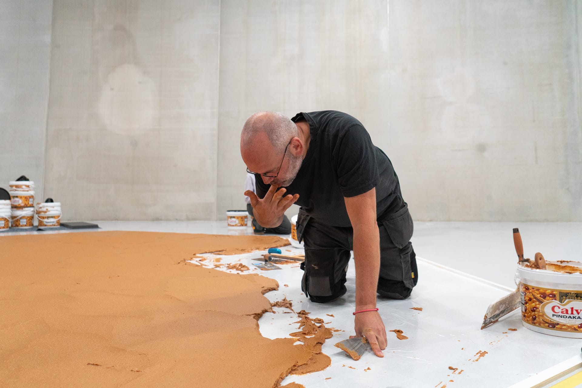 Een man op zijn knieën smeert met een plamuurmes een egale laag pindakaas op een witte vloer in een museumzaal
