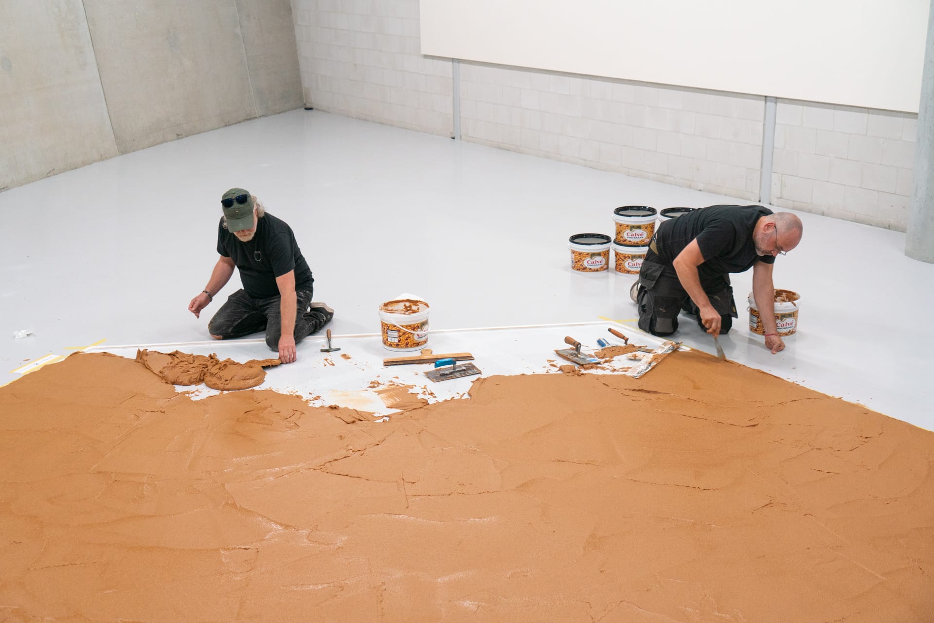 Twee mannen op hun knieën smeren met stukadoorsgereedschap een dikke laag pindakaas uit op een witte museumvloer