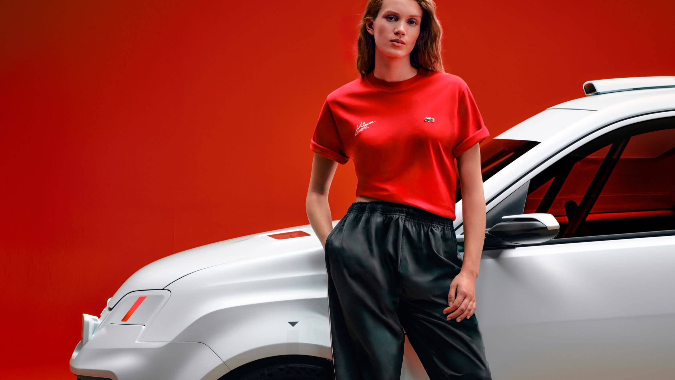Vrouw in een rood Lacoste T-shirt en zwarte broek leunt tegen een witte auto voor een rode achtergrond