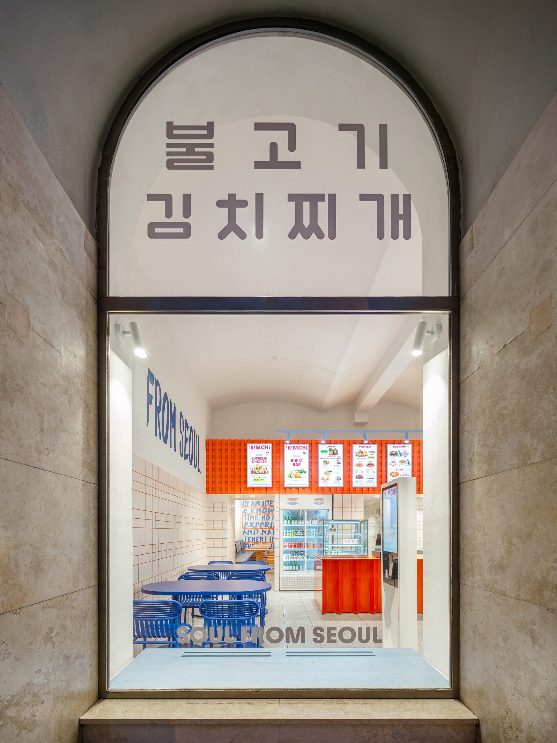 Zicht op een modern Koreaans restaurant door een glazen boogdeur met Koreaanse tekens en blauw meubilair binnen, het gaat om restaurant The Kimchi in Praag