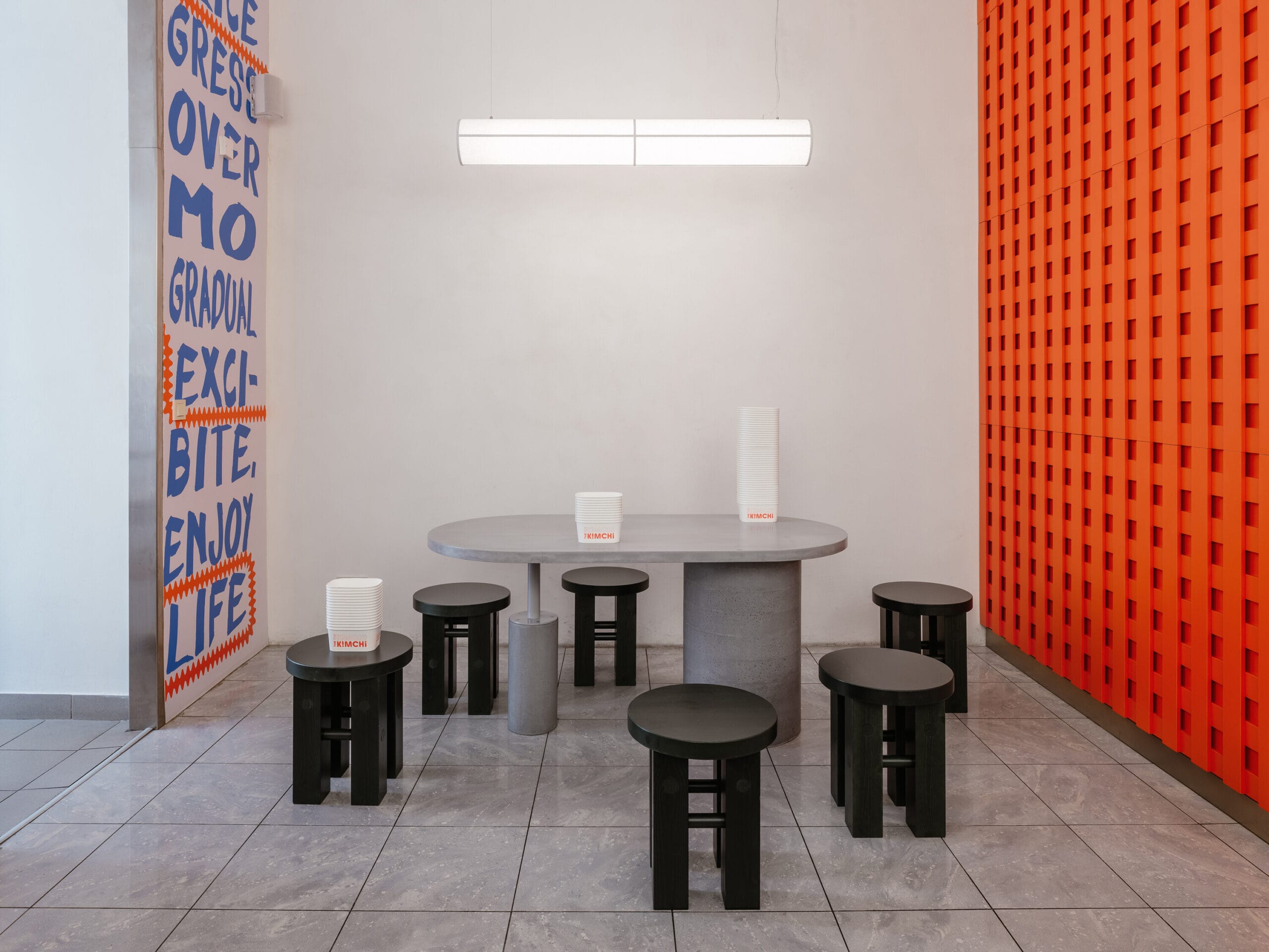 Modern minimalistisch interieur van restaurant The Kimchi in Praag, met een grijze ovale tafel, zwarte krukken en een oranje wand