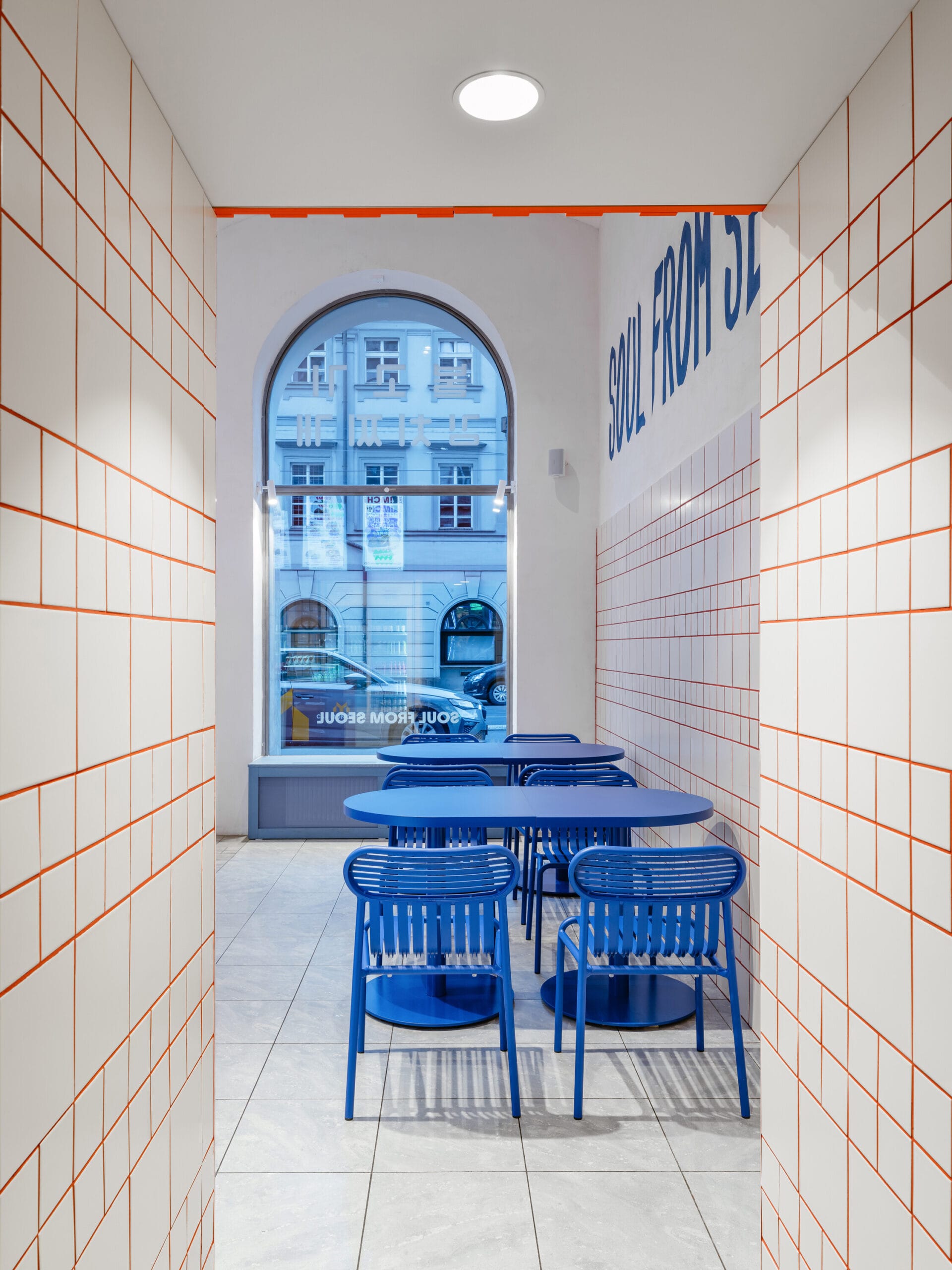 Modern interieur van het restaurant The Kimchi in Praag, met witte tegels, oranje voegen en blauwe tafels en stoelen bij een boogvenster