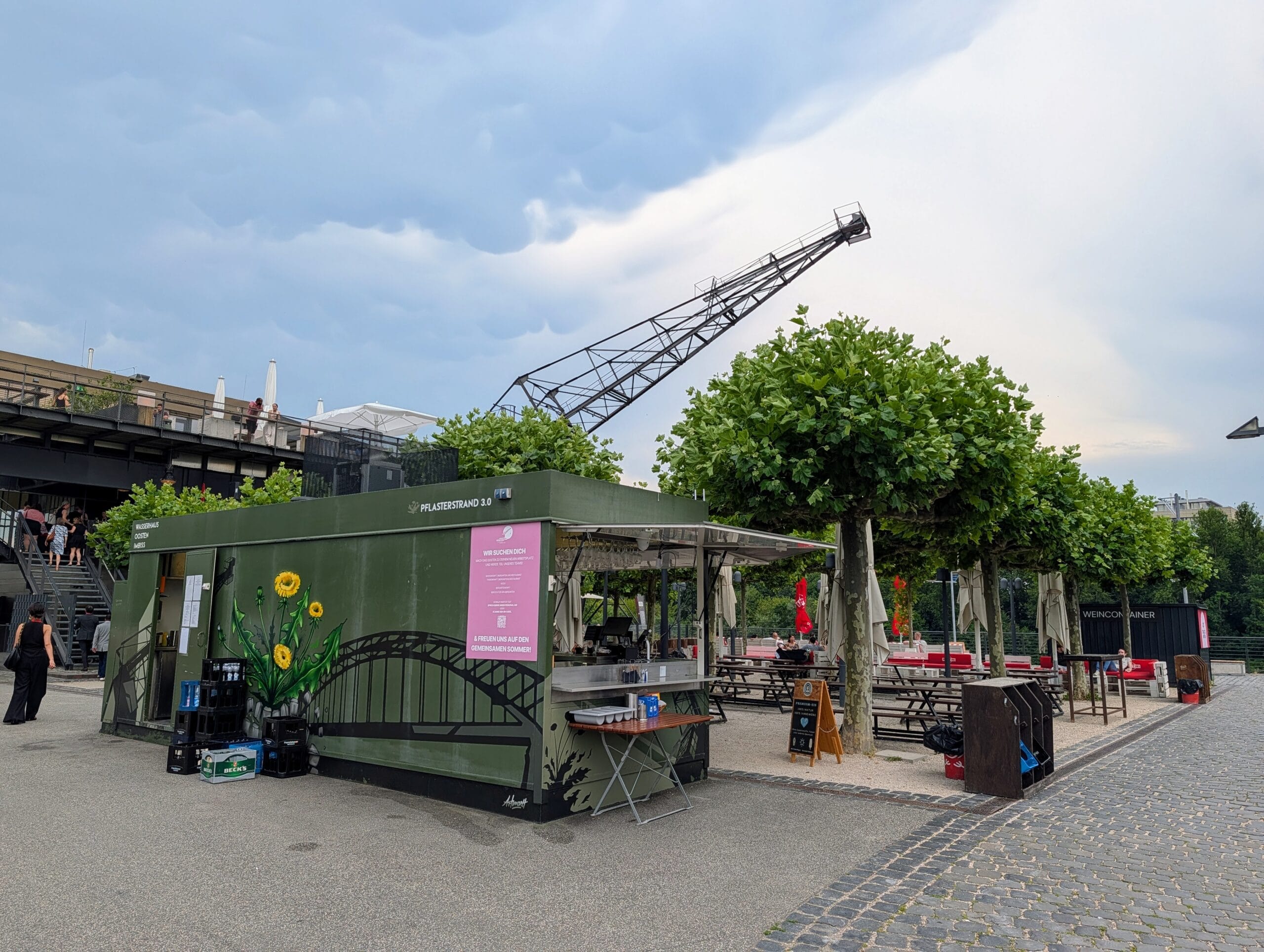 Terras van restaurant OOSTEN met een groene containerbar, bomen en een grote industriële kraan tegen de lucht