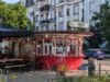 Een rood rond kioskgebouw genaamd Nox Wasserhäuschen met een terras en stickers op de gevel voor een wit appartementencomplex