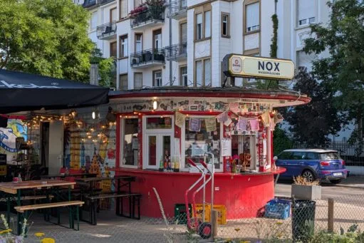 Een rood rond kioskgebouw genaamd Nox Wasserhäuschen met een terras en stickers op de gevel voor een wit appartementencomplex