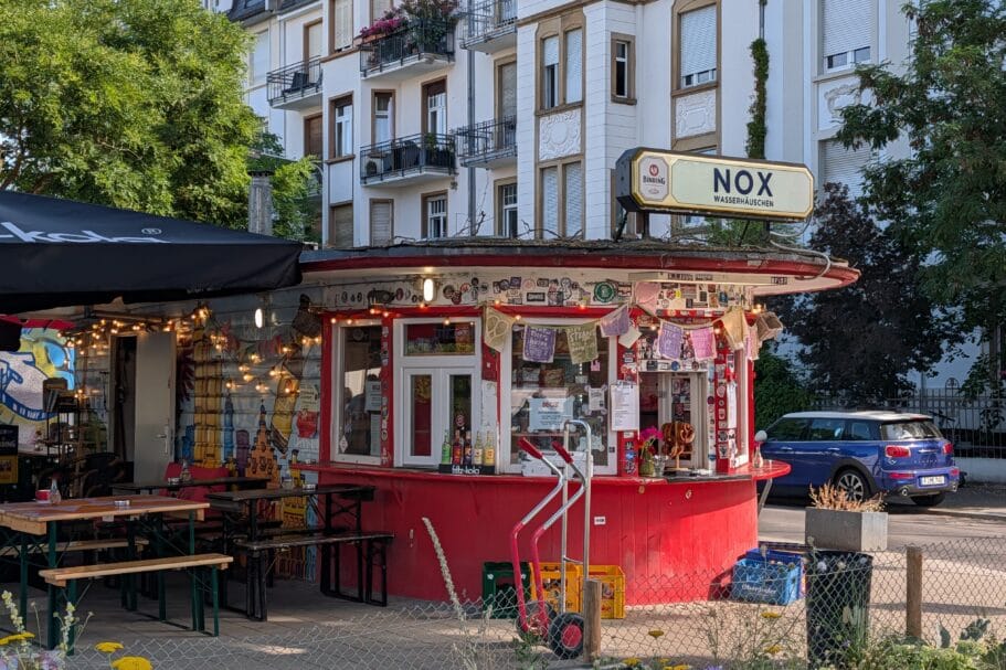 Een rood rond kioskgebouw genaamd Nox Wasserhäuschen met een terras en stickers op de gevel voor een wit appartementencomplex