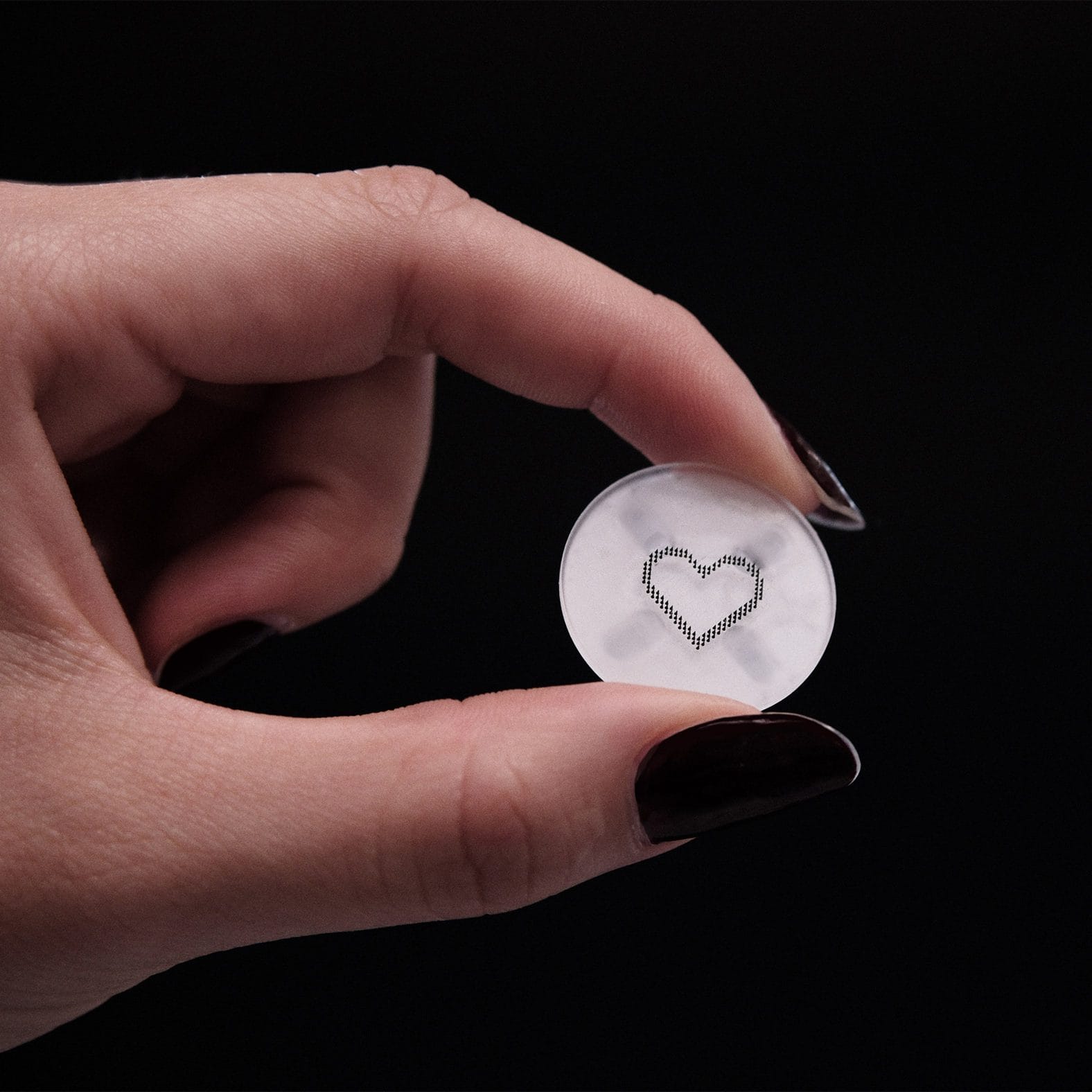 Een hand houdt een kleine, ronde, transparante patch vast met daarop een hartvormig ontwerp van minuscule naaldjes