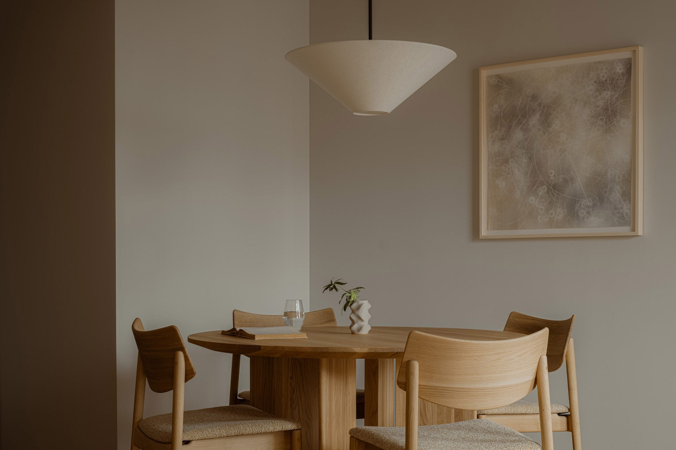Minimalistisch interieur met een ronde houten eettafel, vier bijpassende stoelen, een hanglamp en abstracte kunst