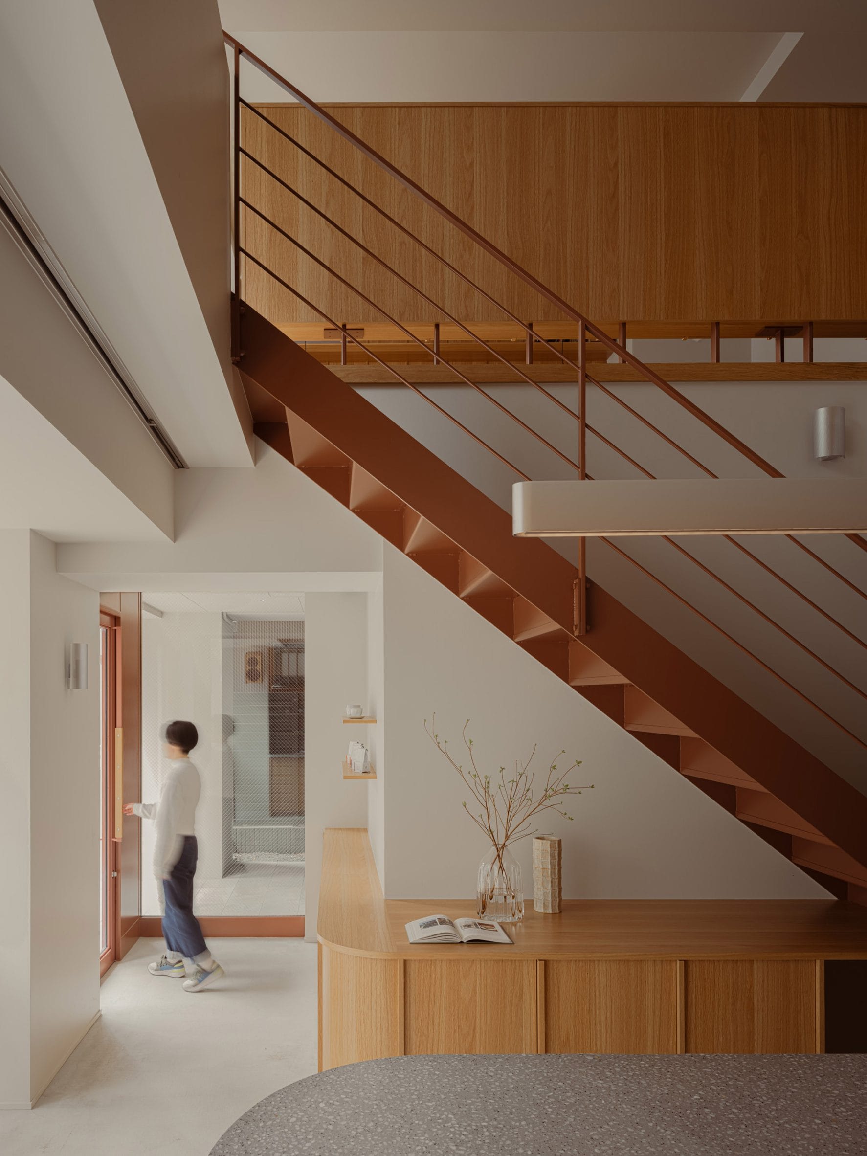Modern interieur met een roestbruine stalen trap, houten kasten en een persoon die door een glazen deur loopt
