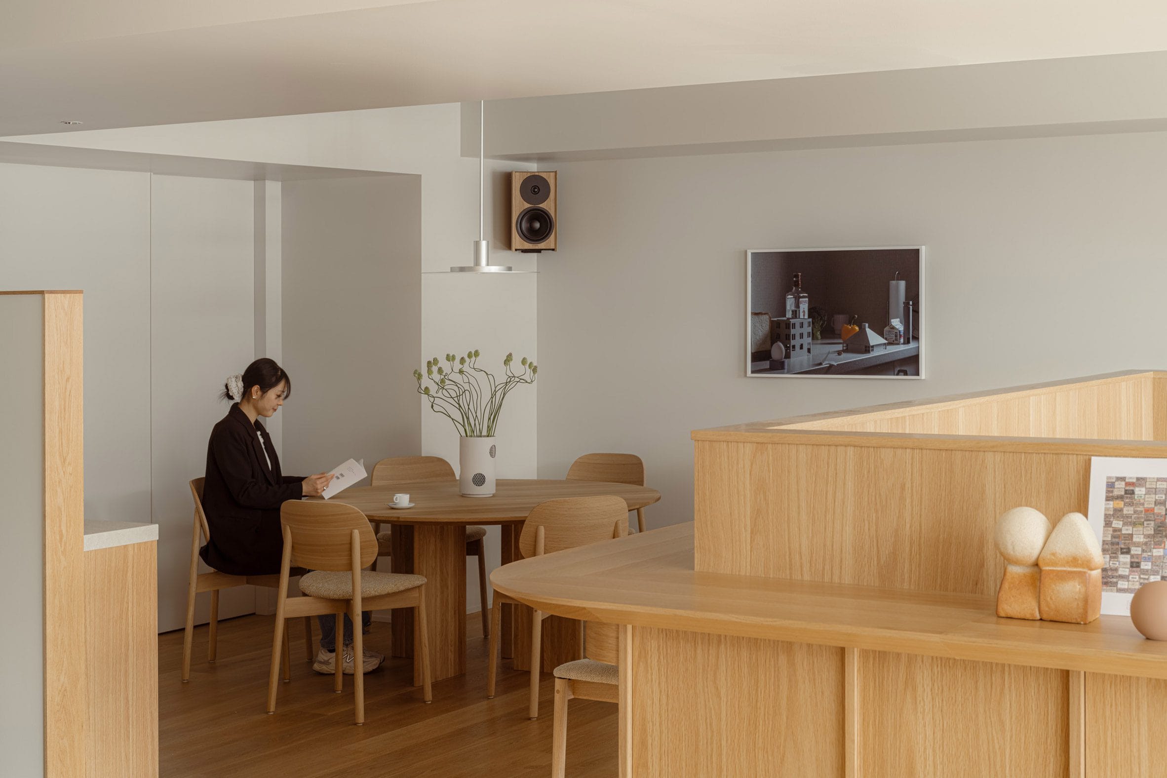 Een vrouw leest een boek aan een ronde houten eettafel in een minimalistisch, licht interieur met houten accenten