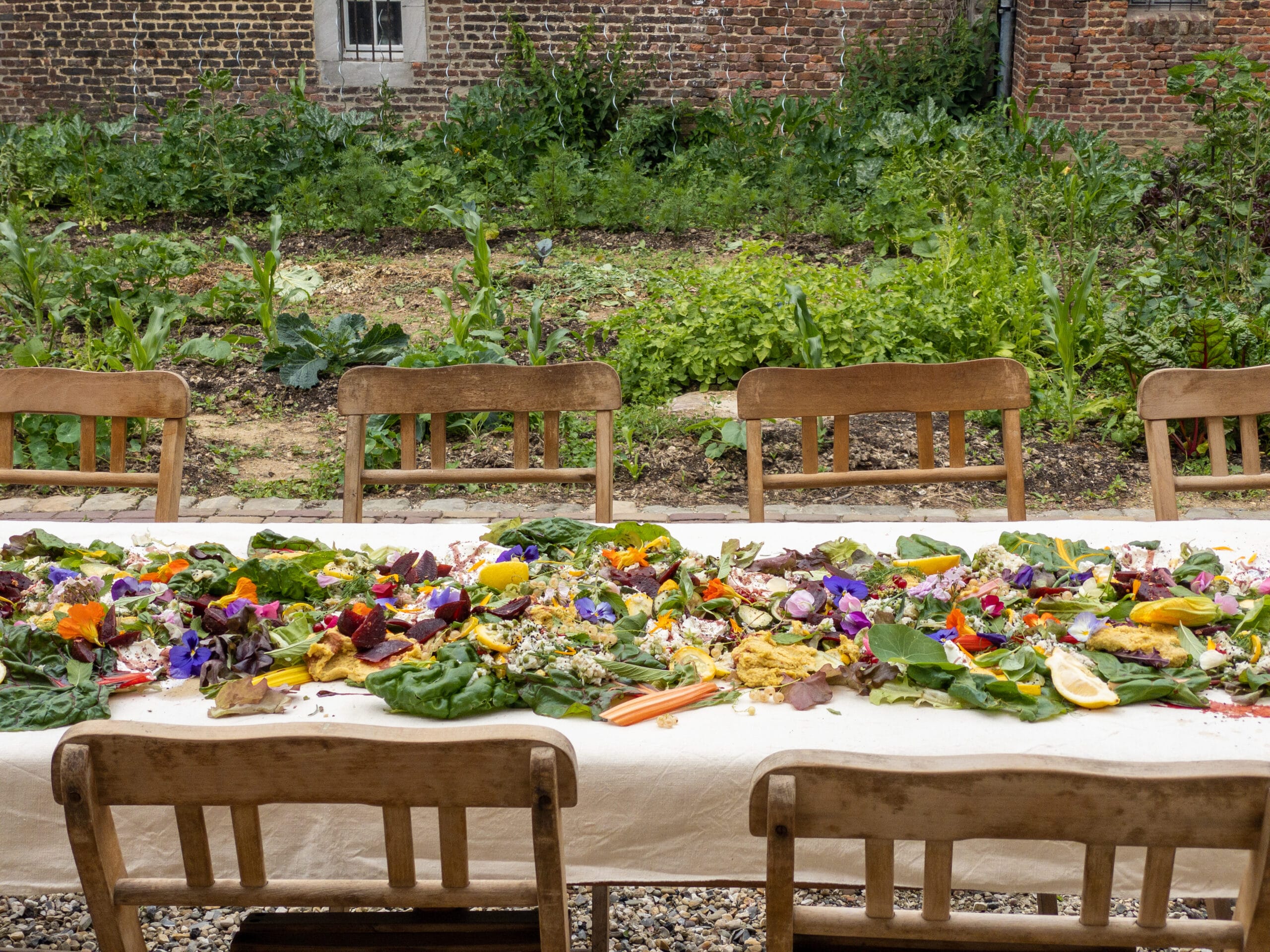 Een lange tafel met wit linnen kleed vol kleurrijke groenten, kruiden en bloemen, met houten stoelen in een tuin