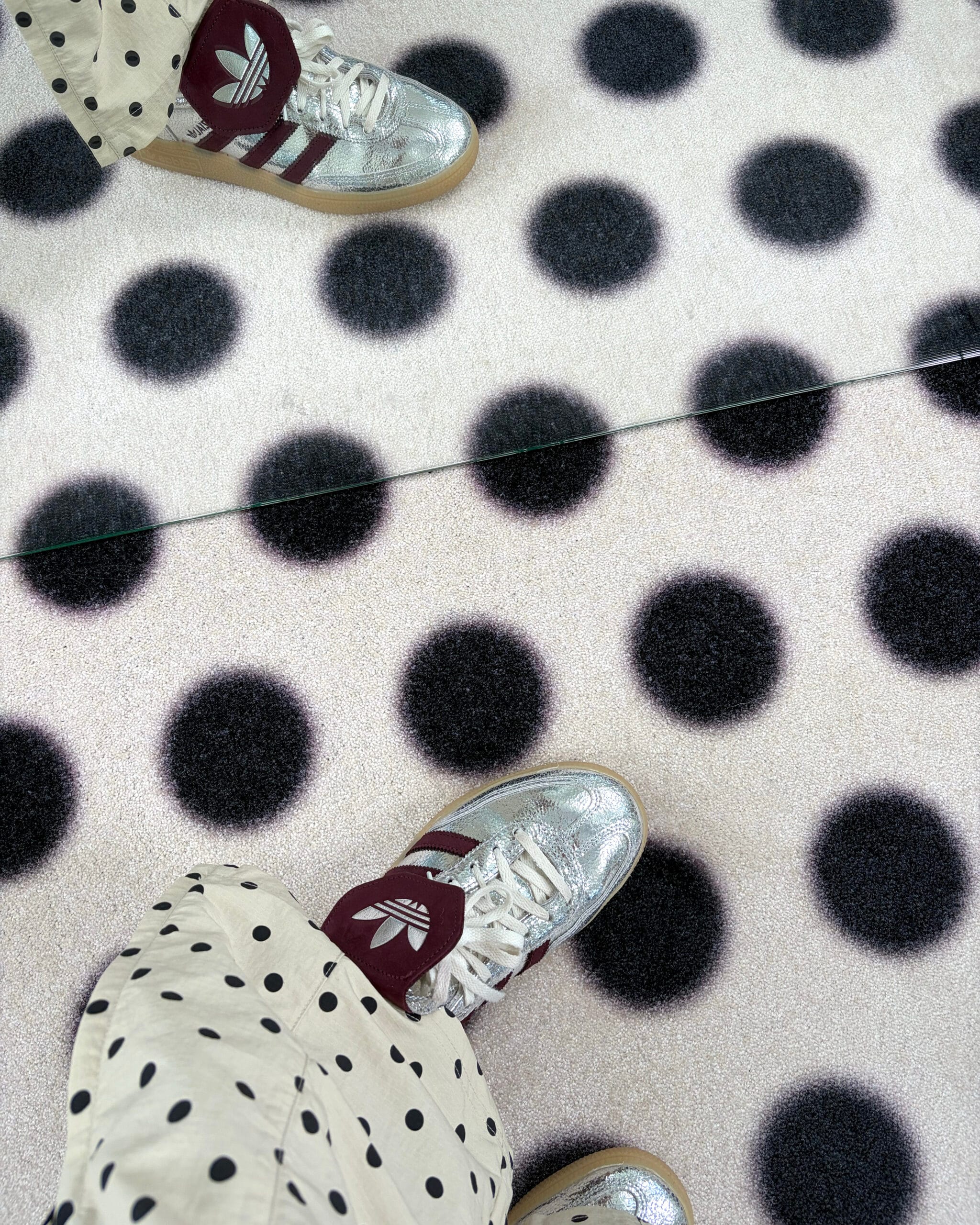 Bovenaanzicht van een witte Maison Deux deurmat met grote zwarte stippen, waarop iemand met zilveren sneakers staat