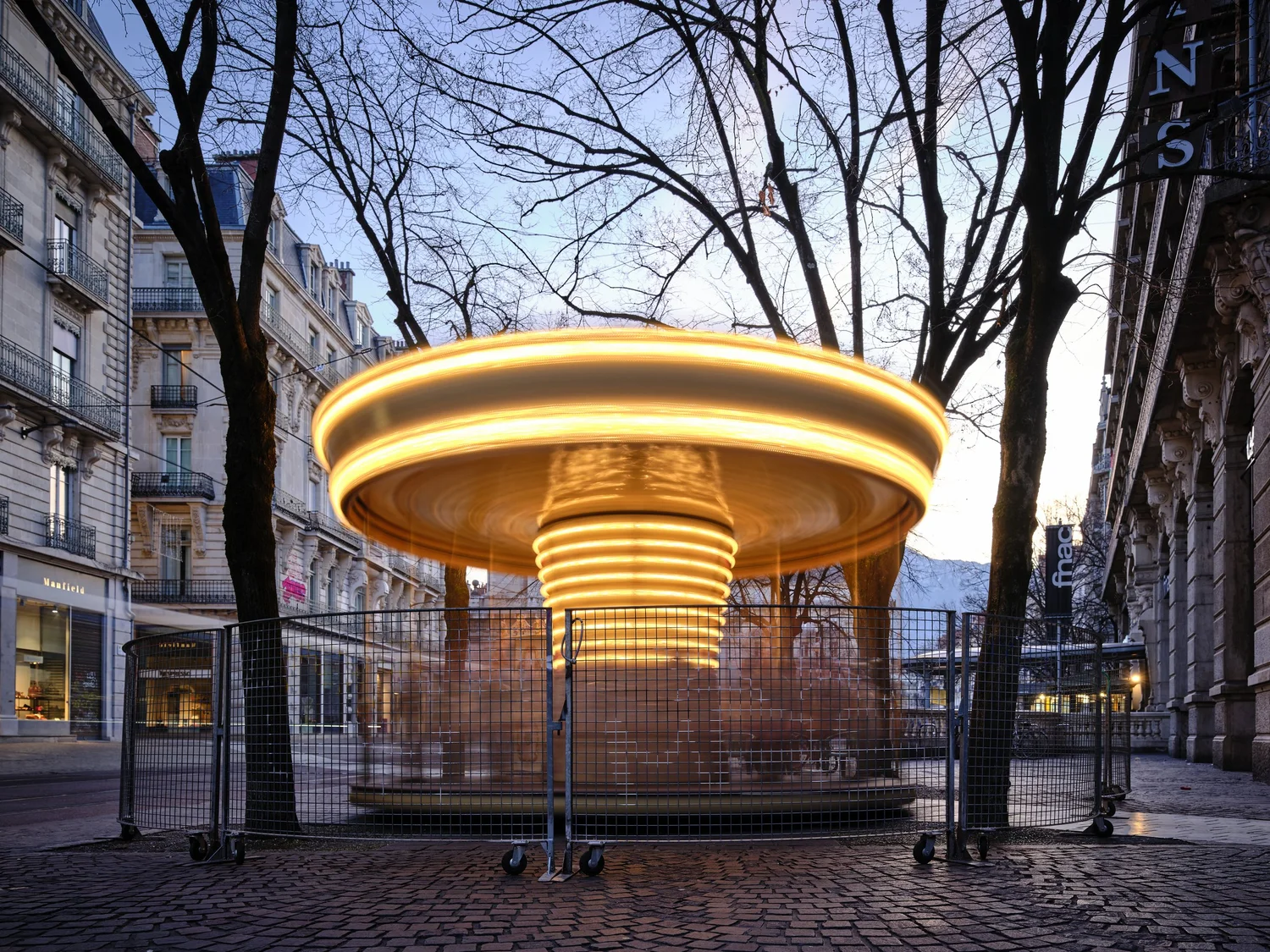 Draaiende, geel verlichte carrousel vastgelegd met lange sluitertijd op een stadsplein tussen bomen en gebouwen