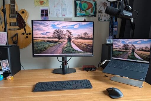 Werkplek met een houten bureau, AOC-monitor, laptop op een standaard, draadloos toetsenbord, muis en een microfoon