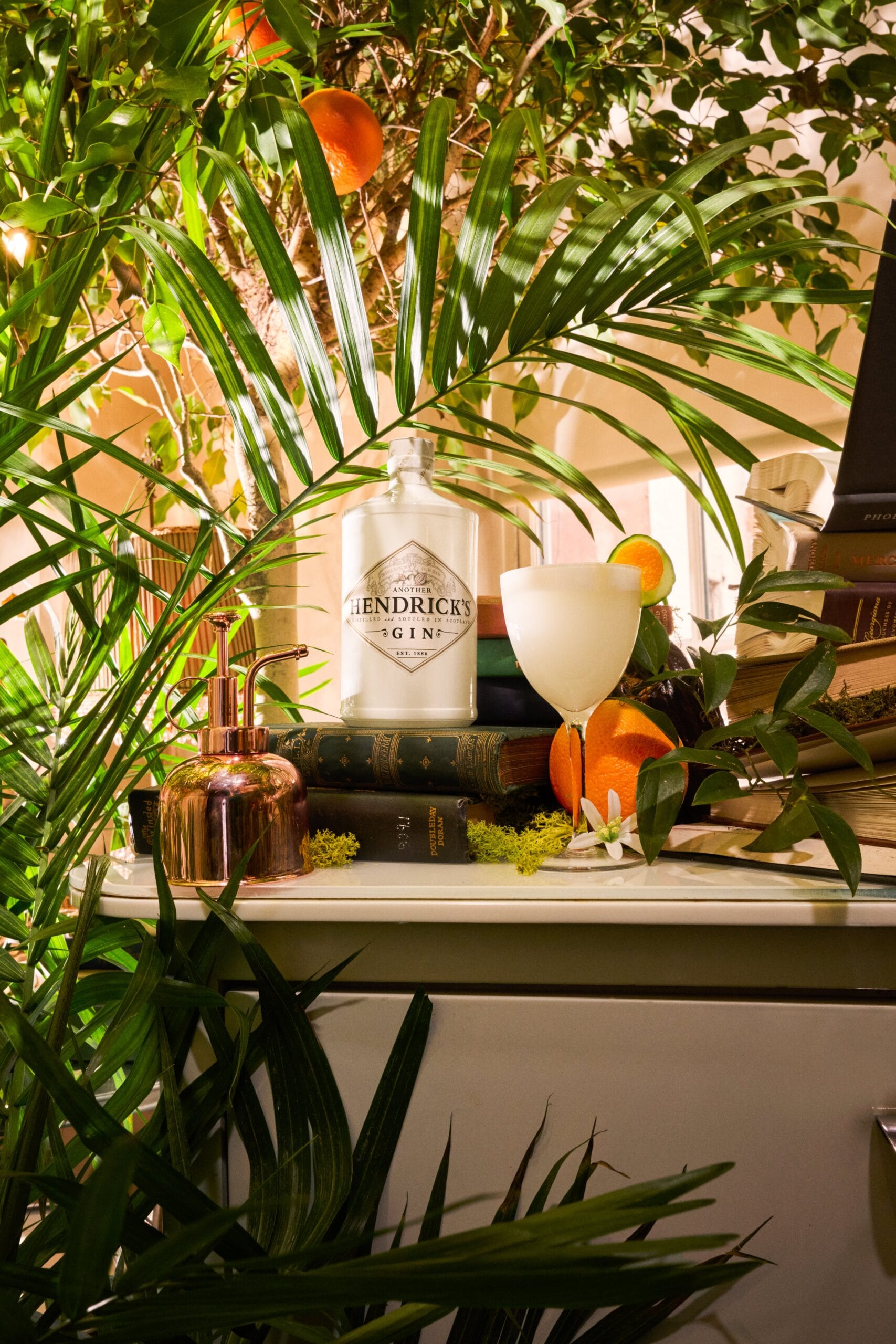 De witte fles Another Hendrick’s Gin staat op een stapel boeken naast een cocktailglas in een tropische setting