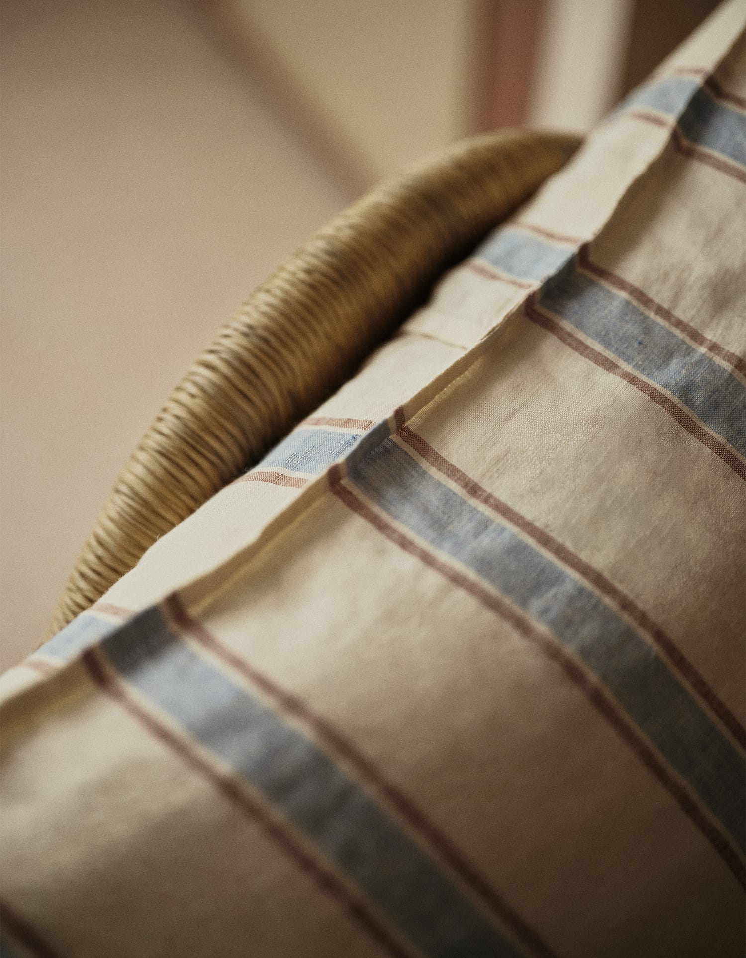 Close-up van een beige kussen met blauwe en bruine strepen tegen de leuning van een rotan stoel, uit de Essentials collectie van Bonsoirs