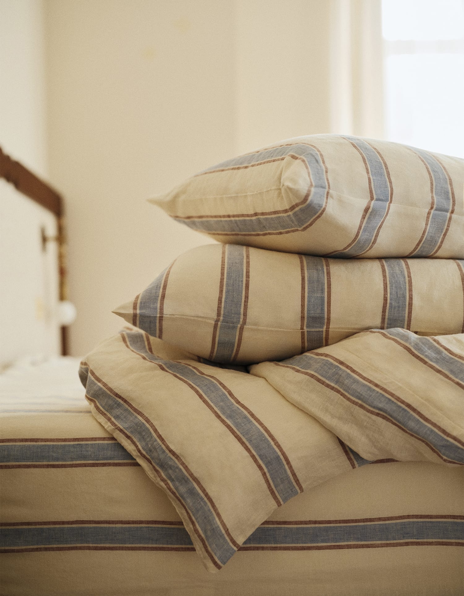 Stapel beige kussens en een dekbed met blauwe en bruine strepen op een bed, uit de Essentials collectie van Bonsoirs