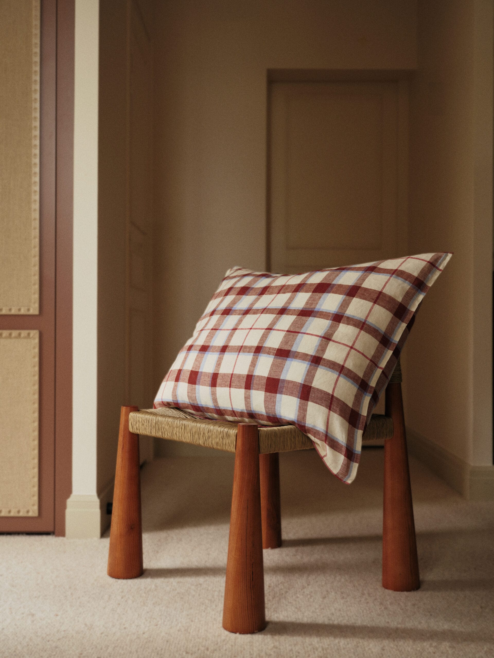 Vierkant kussen met een rood, blauw en beige ruitpatroon op een houten krukje met een zitting van gevlochten touw, uit de Essentials collectie van Bonsoirs